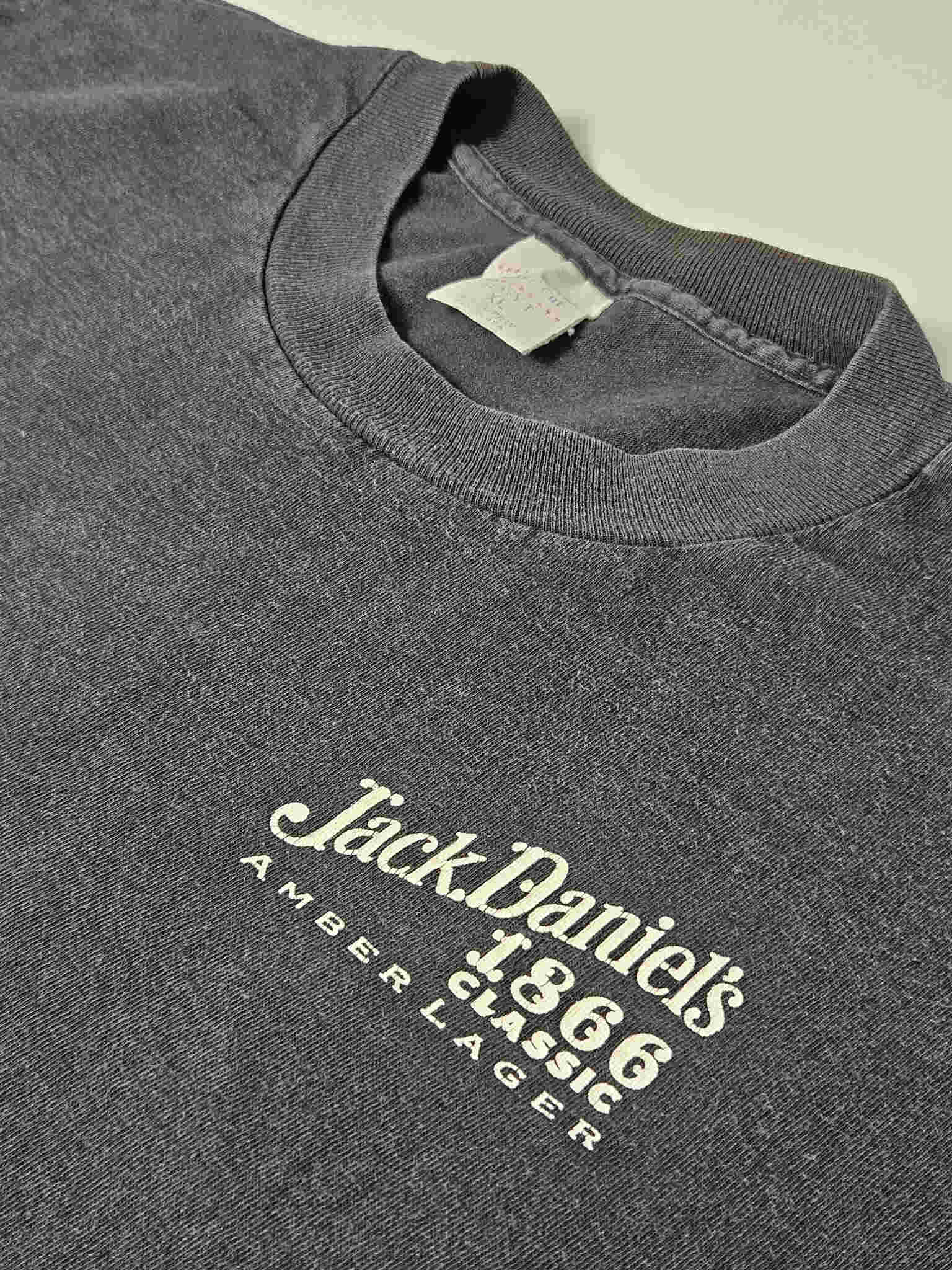 "Close-up van het witte 'Jack Daniel's 1866 Classic Amber Lager' logo op de linkerborst van een zwart vintage T-shirt. De details van de print en de subtiele textuur van de stof zijn duidelijk zichtbaar, wat de authenticiteit van dit tijdloze slow fashion item onderstreept."