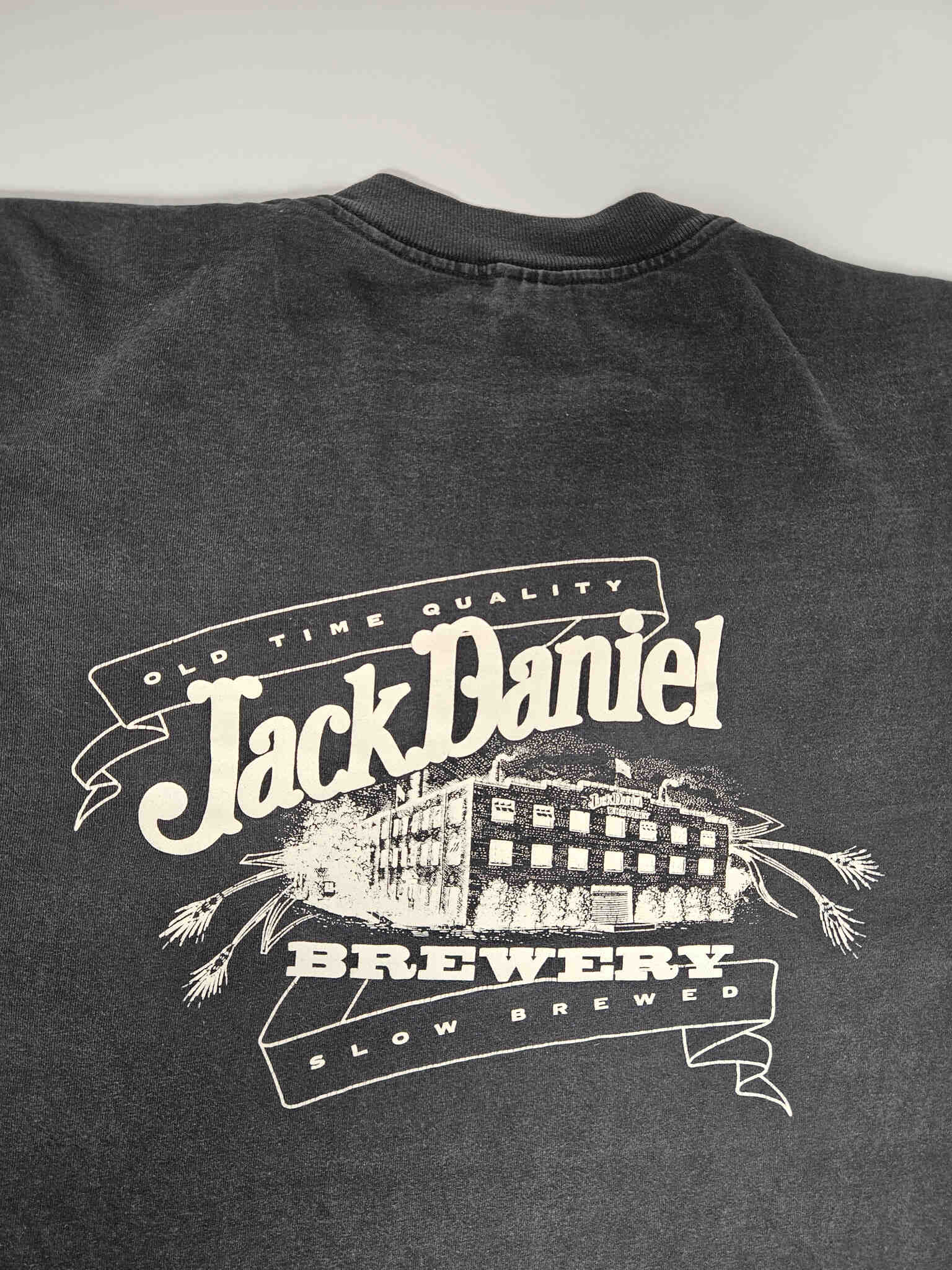 "Gedetailleerde close-up van de grote witte print op de achterkant van een zwart vintage T-shirt. De print toont 'OLD TIME QUALITY Jack Daniel's BREWERY SLOW BREWED' met een illustratie van een brouwerijgebouw en graanaren. Deze unieke grafische weergave draagt bij aan de zeldzaamheid van dit slow fashion verzamelstuk."