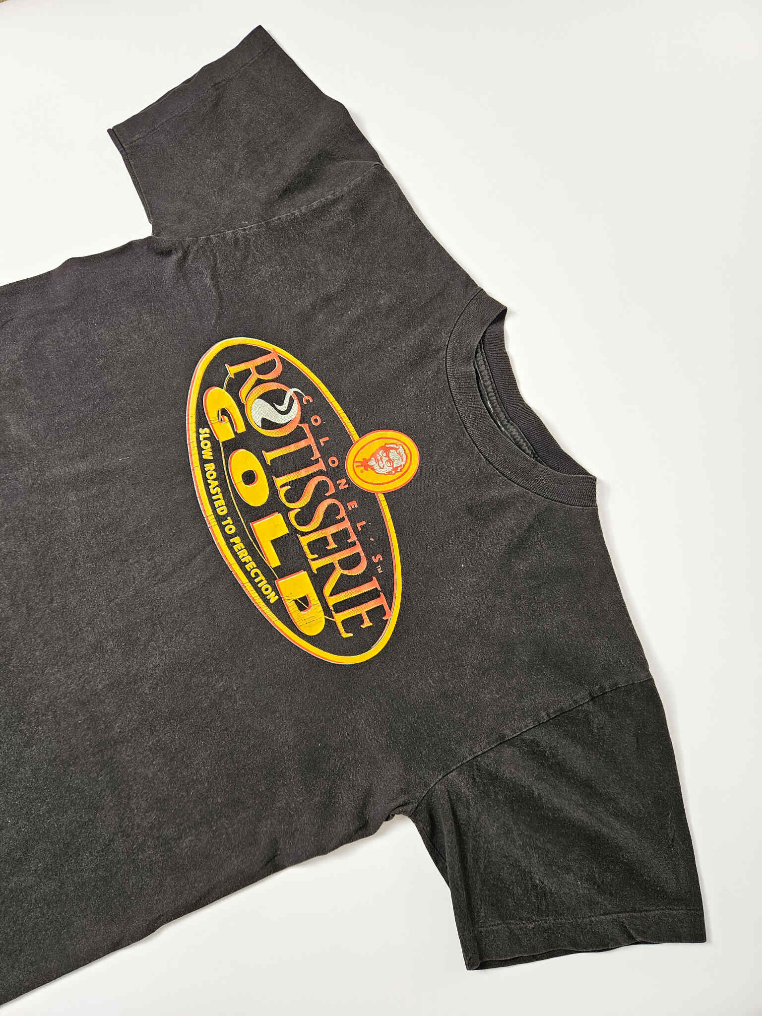 "Schuin perspectief van een zwart vintage T-shirt, waarop het grote 'COLONEL'S ROTISSERIE GOLD' logo met het Colonel Sanders portret te zien is. De textuur van de stof en de afwerking van de print zijn duidelijk, wat de kwaliteit en de 'pre-loved' status van dit duurzame kledingstuk accentueert."