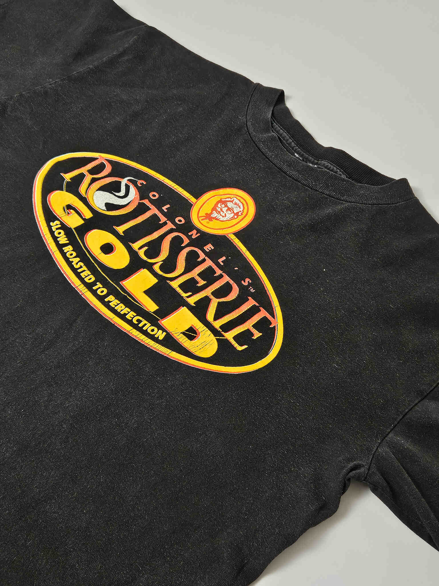 "Schuin perspectief van een zwart vintage T-shirt, waarop het grote 'COLONEL'S ROTISSERIE GOLD' logo met het Colonel Sanders portret te zien is. De textuur van de stof en de afwerking van de print zijn duidelijk, wat de kwaliteit en de 'pre-loved' status van dit duurzame kledingstuk accentueert."