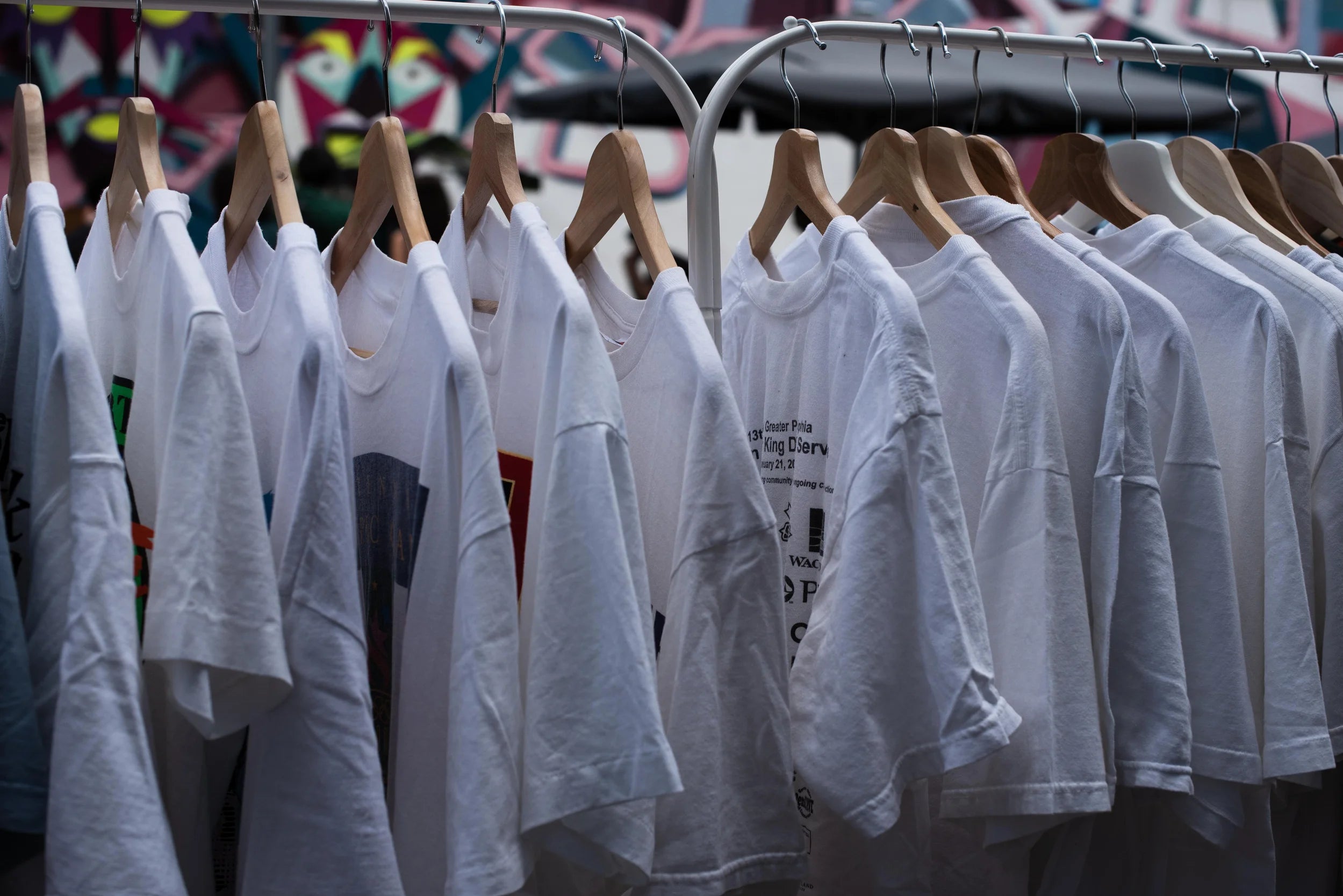 Rij met authentieke witte vintage T-shirts uit de jaren ’80 en ’90 aan houten hangers op een kledingrek – onder andere Hanes, Fruit of the Loom en screen printed bandshirts – gefotografeerd in daglicht tijdens een vintage kledingmarkt, met een kleurrijke graffiti-achtergrond.