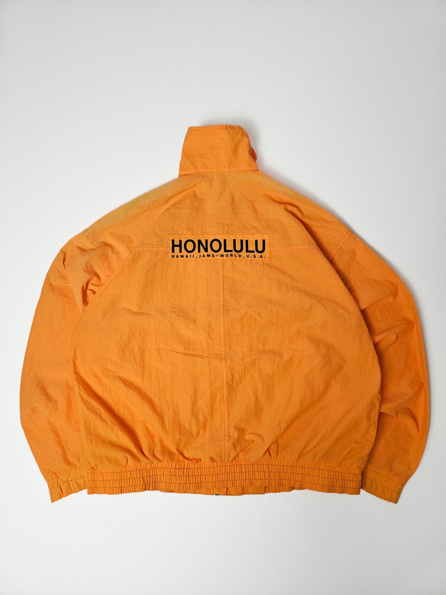 Achterkant van de oranje windbreaker met grote zwarte print: “HONOLULU – Hawaii, Jams World, U.S.A.”