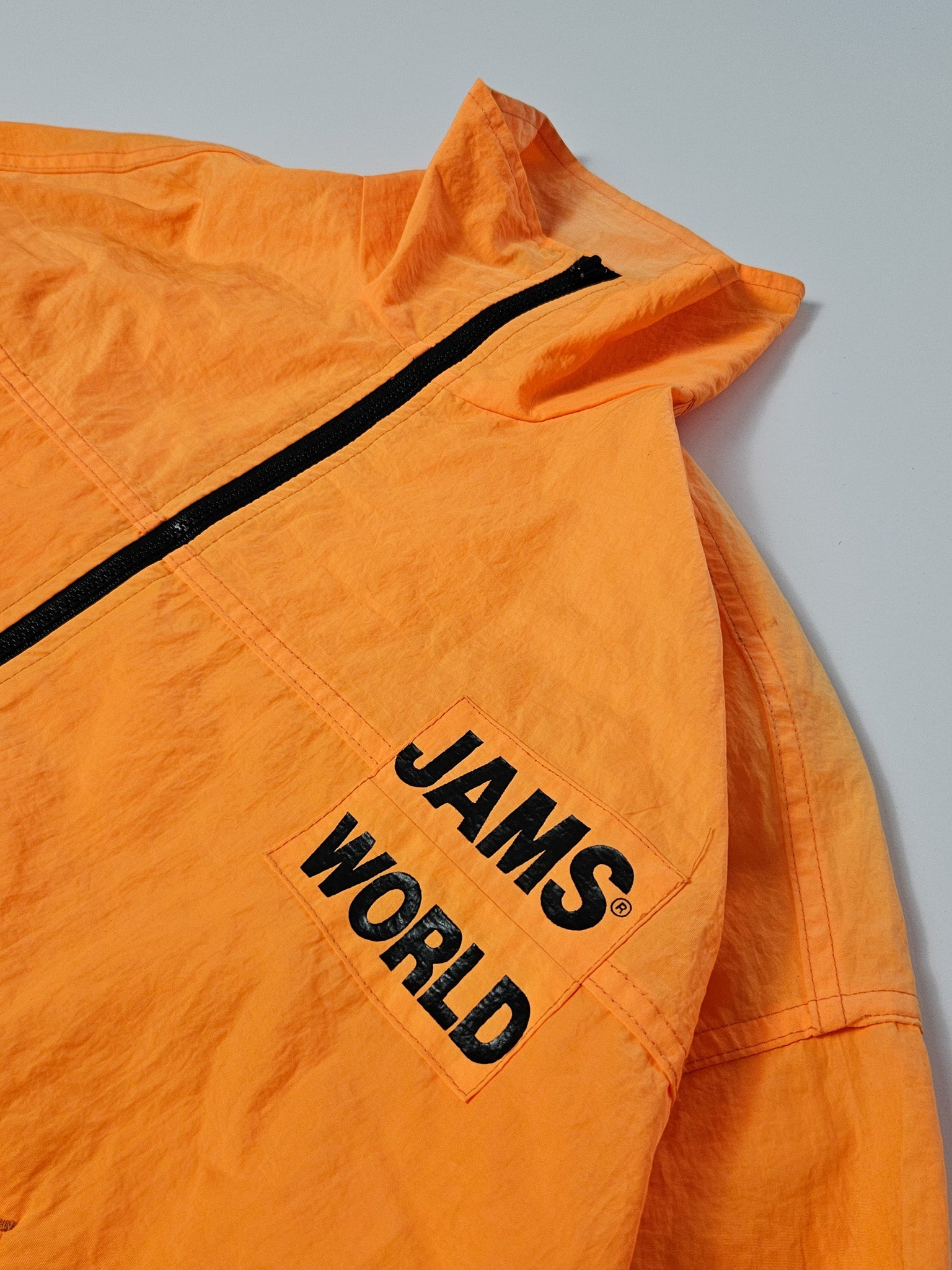 Detail van de zwarte JAMS WORLD print op de borst van de oranje windbreaker