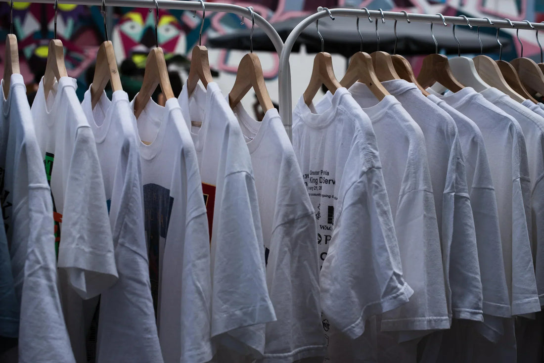 “Rij met authentieke witte vintage T-shirts uit de jaren ’80 en ’90 aan houten hangers op een kledingrek – onder andere Hanes, Fruit of the Loom en screen printed bandshirts – gefotografeerd in daglicht tijdens een vintage kledingmarkt