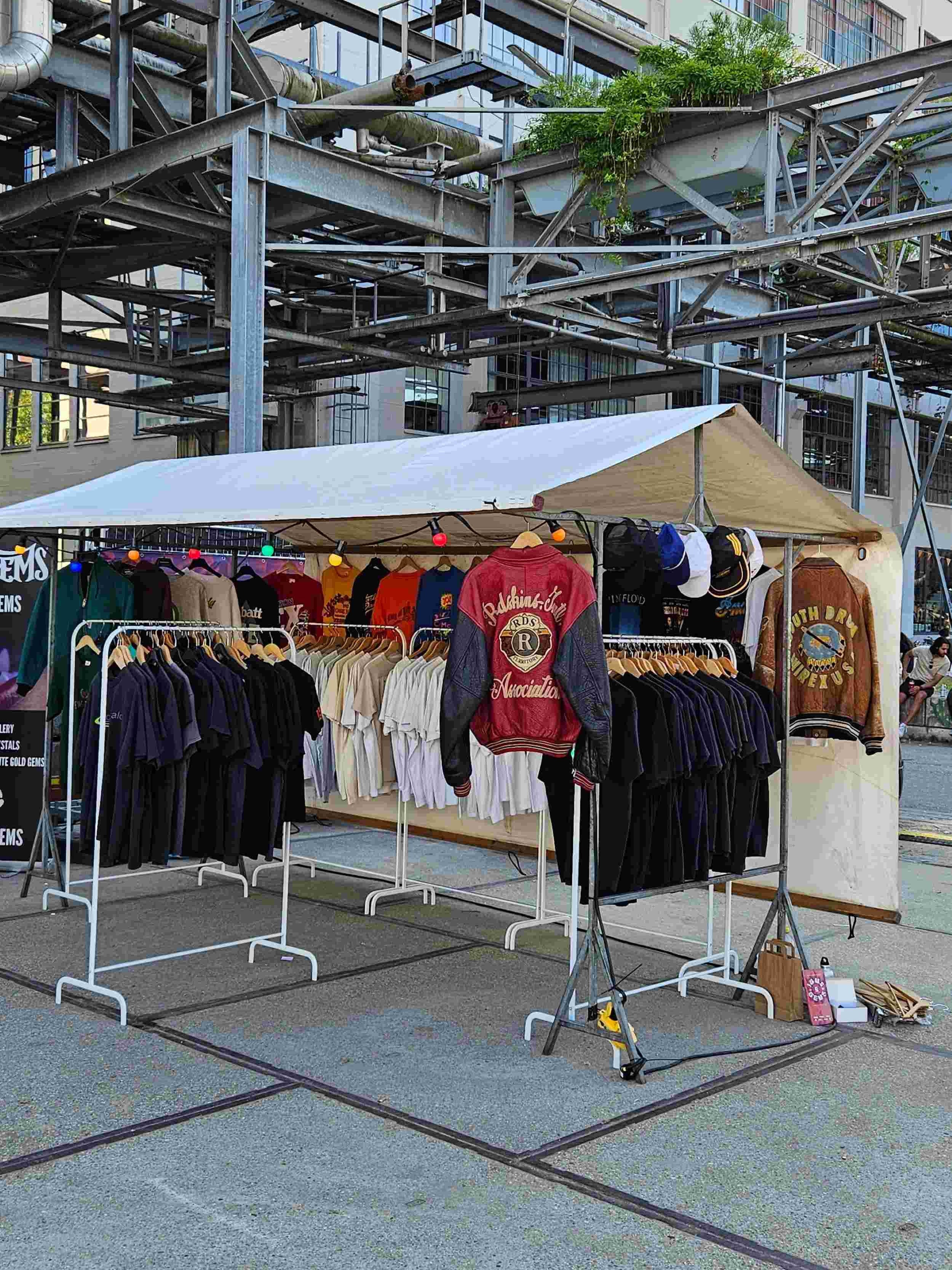 Vintage kledingkraam op een industriële marktlocatie, met rekken vol T-shirts, varsity jacks, caps en kleurrijke lichtsnoeren onder een beige tentdoek – Slow Fashion Vintage pop-up.