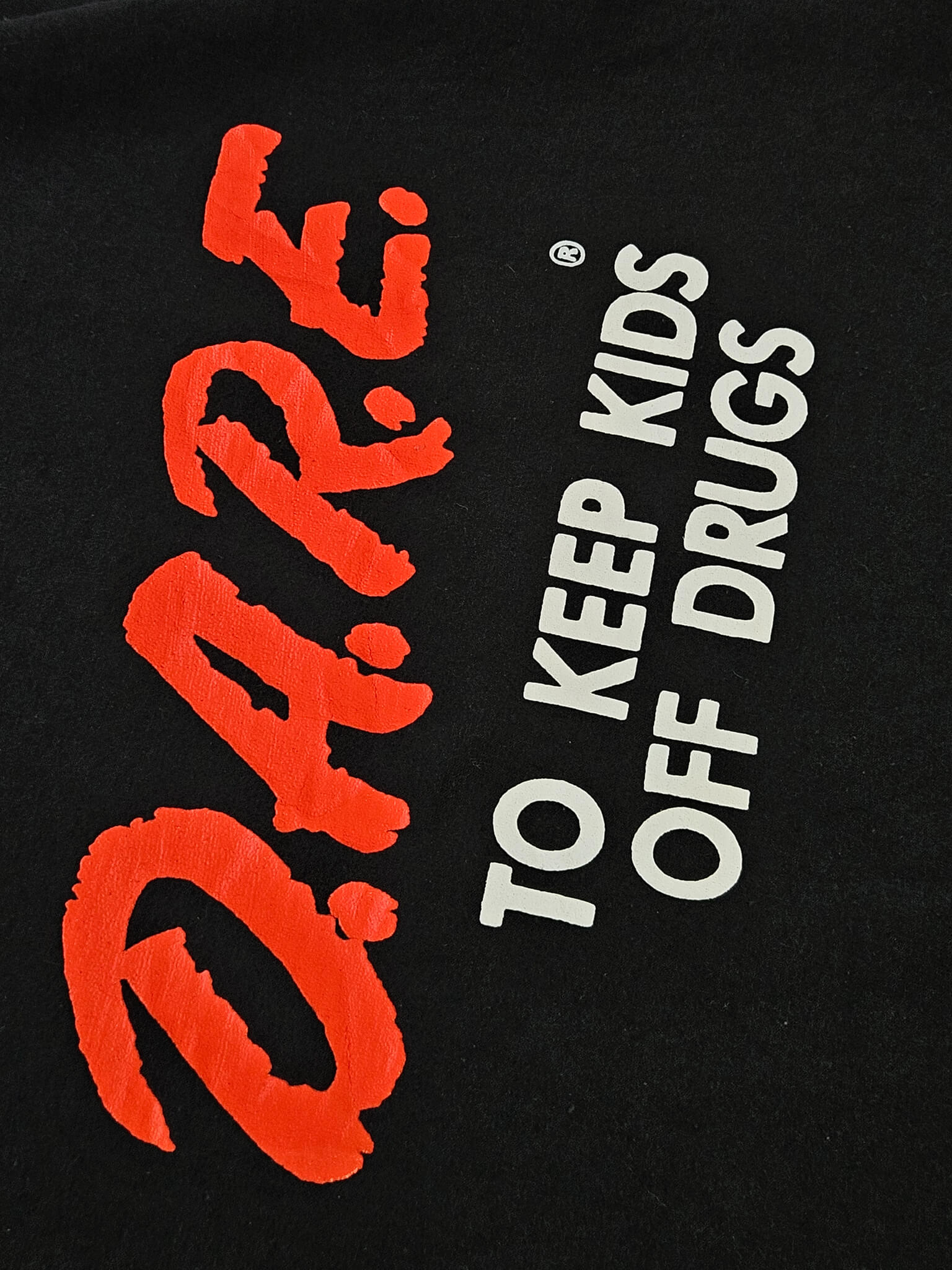 ⦁ Detail: Close-up rood D.A.R.E. brushscript-logo en witte "To Keep Kids Off Drugs" tekst op zwarte stof
