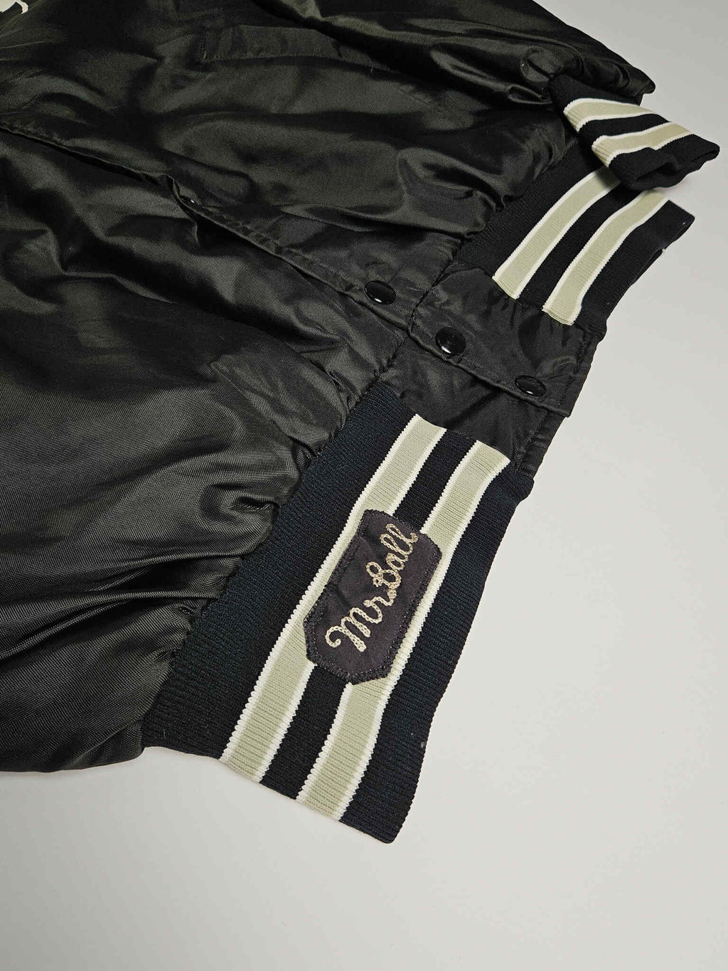 "Close-up van de heup van een zwart vintage satijnen bomberjack, met een geborduurde 'Mr. Ball' patch op de geribbelde manchet met crèmekleurige en zwarte strepen. Dit detail benadrukt de unieke, handgemaakte elementen die kenmerkend zijn voor slow fashion vintage kledingstukken."

