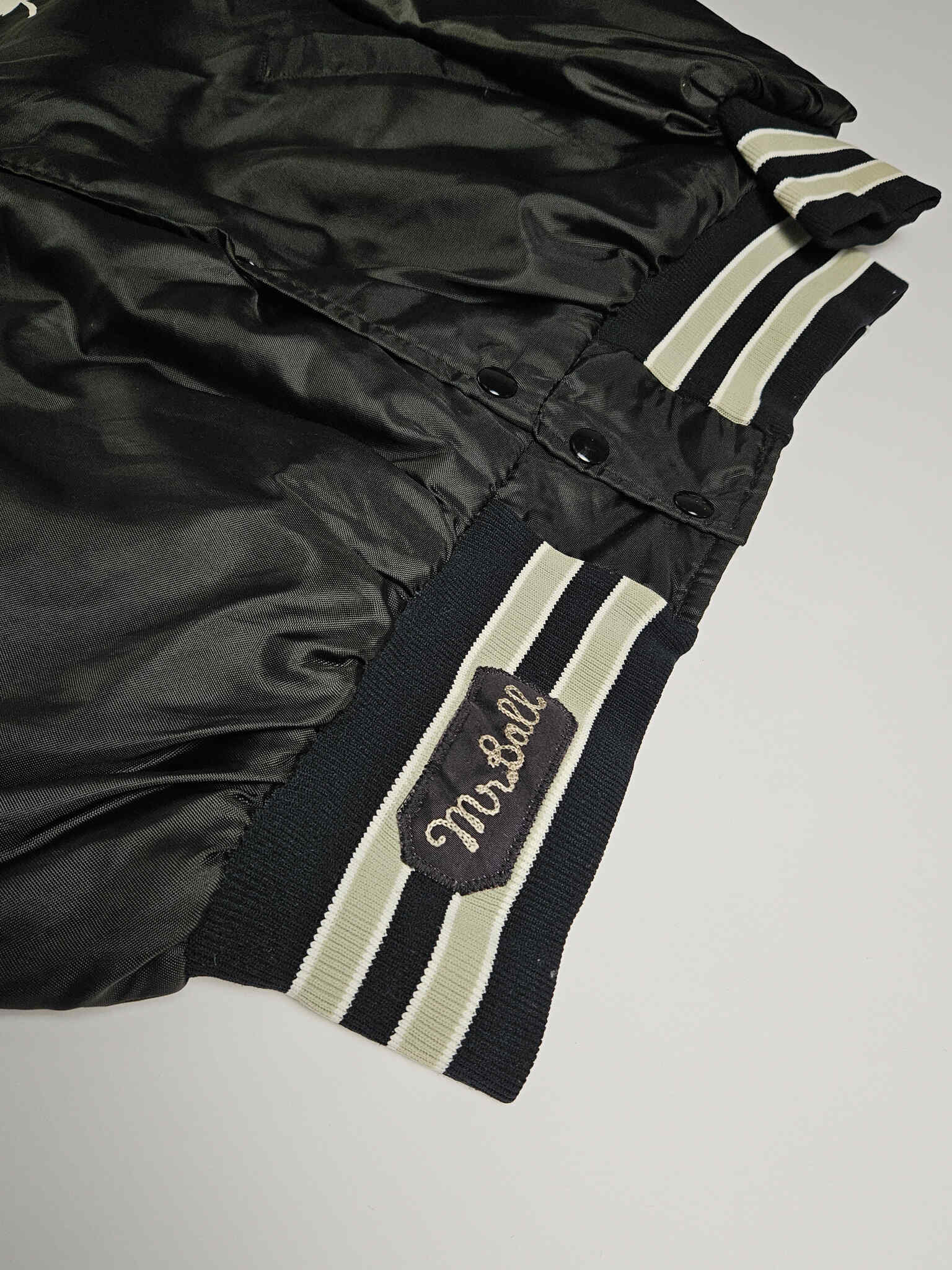 "Close-up van de heup van een zwart vintage satijnen bomberjack, met een geborduurde 'Mr. Ball' patch op de geribbelde manchet met crèmekleurige en zwarte strepen. Dit detail benadrukt de unieke, handgemaakte elementen die kenmerkend zijn voor slow fashion vintage kledingstukken."

