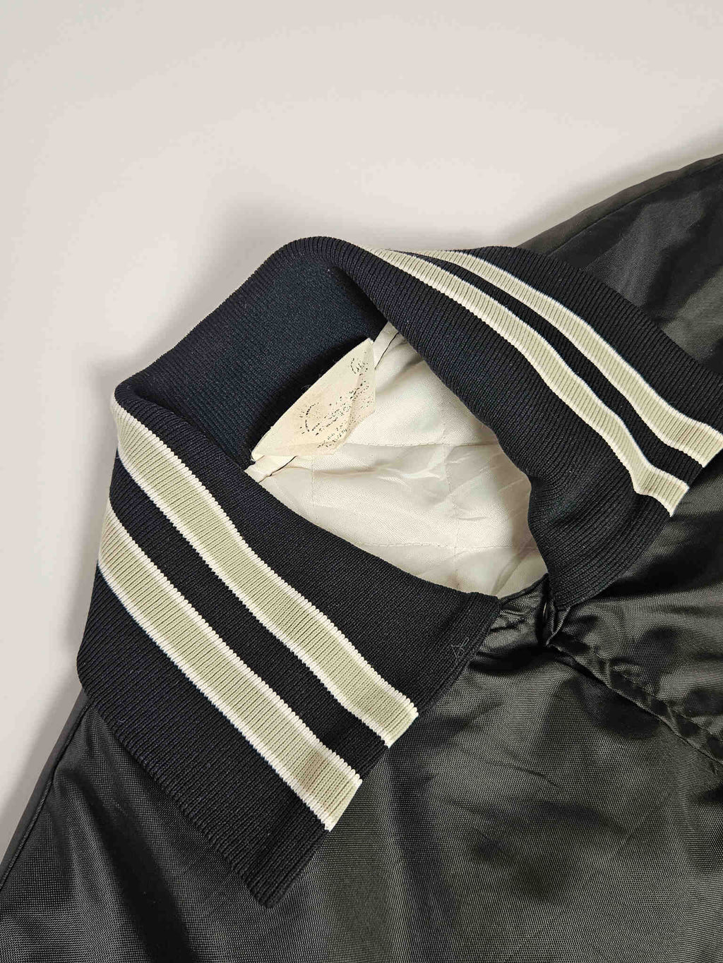 "Close-up van de geribbelde kraag van een zwart vintage satijnen bomberjack, met opvallende crèmekleurige en zwarte strepen. De witte gewatteerde voering is zichtbaar aan de binnenkant. Dit detail toont de kwaliteit en het tijdloze design van dit duurzame slow fashion kledingstuk."

