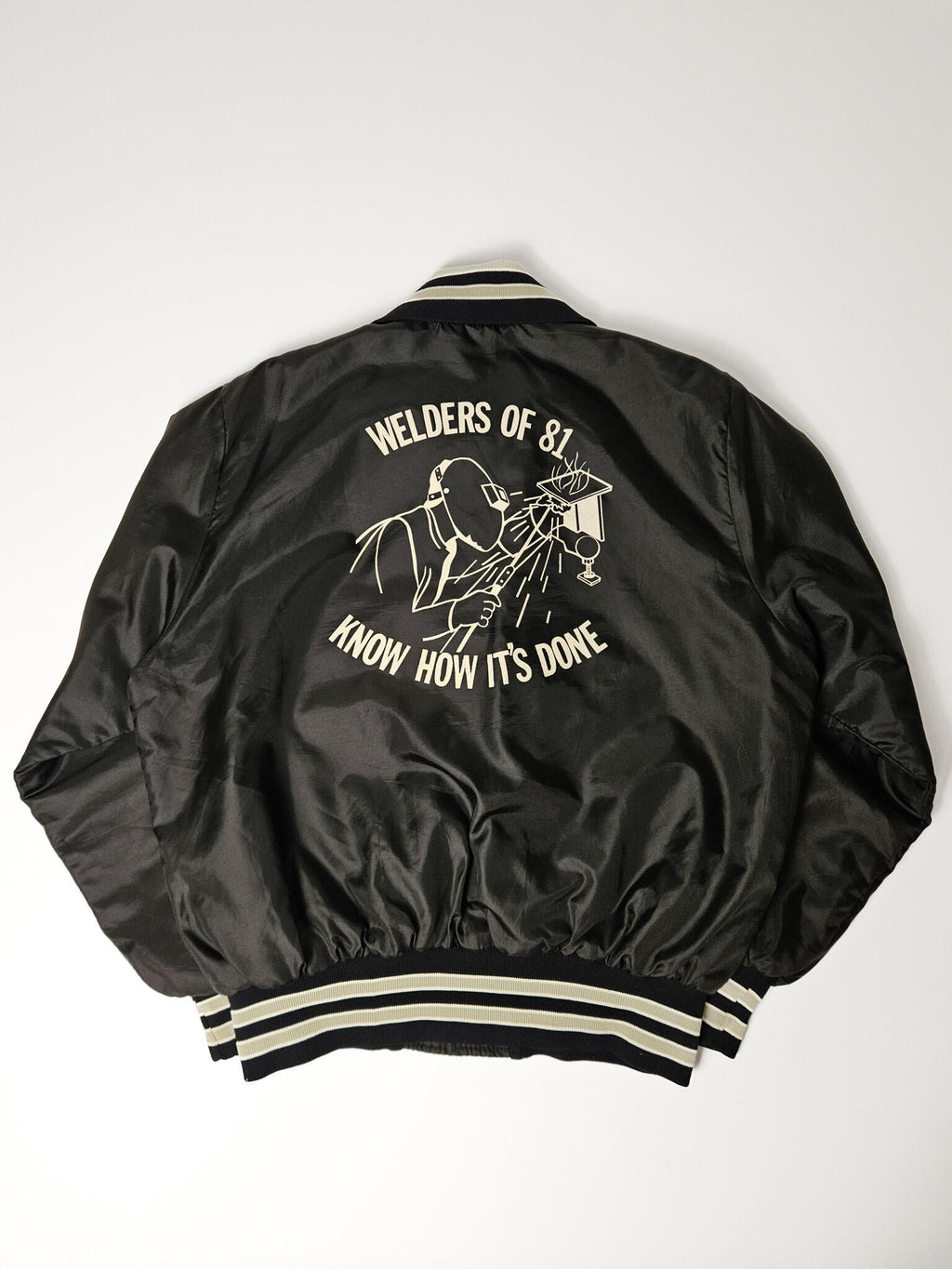 "Achterkant van een zwart vintage satijnen bomberjack met een opvallende witte grafische print van een lasser en de tekst 'Welders of 81, Know How It's Done'. De geribbelde zoom met crèmekleurige en zwarte strepen is zichtbaar. Dit is een uniek stuk dat de duurzaamheid en het verhaal van slow fashion vintage vertegenwoordigt."

