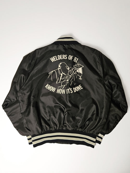 "Achterkant van een zwart vintage satijnen bomberjack met een opvallende witte grafische print van een lasser en de tekst 'Welders of 81, Know How It's Done'. De geribbelde zoom met crèmekleurige en zwarte strepen is zichtbaar. Dit is een uniek stuk dat de duurzaamheid en het verhaal van slow fashion vintage vertegenwoordigt."

