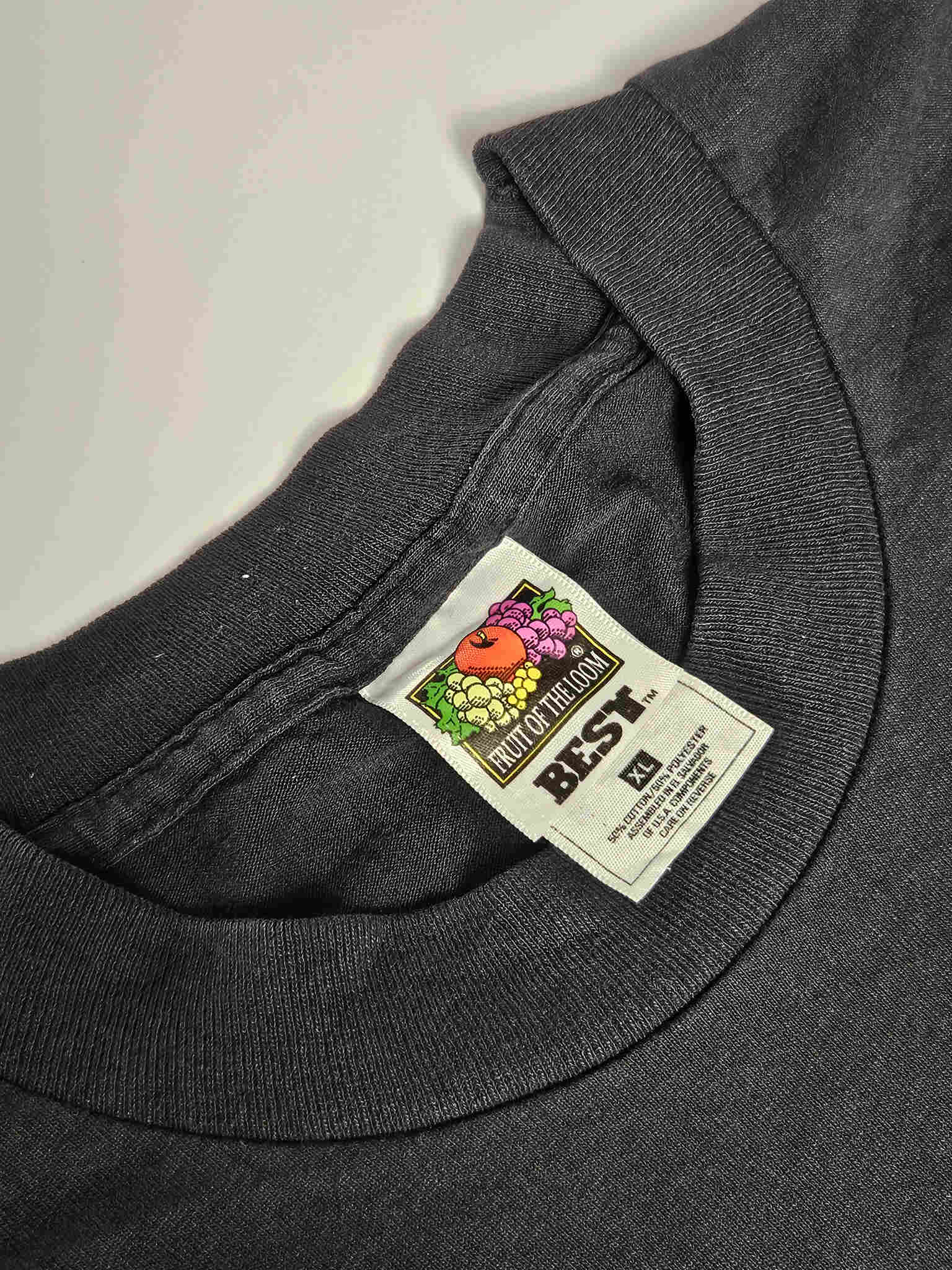"Close-up van het label in de nek van een zwart vintage T-shirt, waarop 'Fruit of the Loom BEST XL' staat. Het label toont de klassieke Fruit of the Loom fruitafbeelding. Dit helpt bij het authentificeren van de leeftijd en het merk van dit duurzame slow fashion T-shirt."










