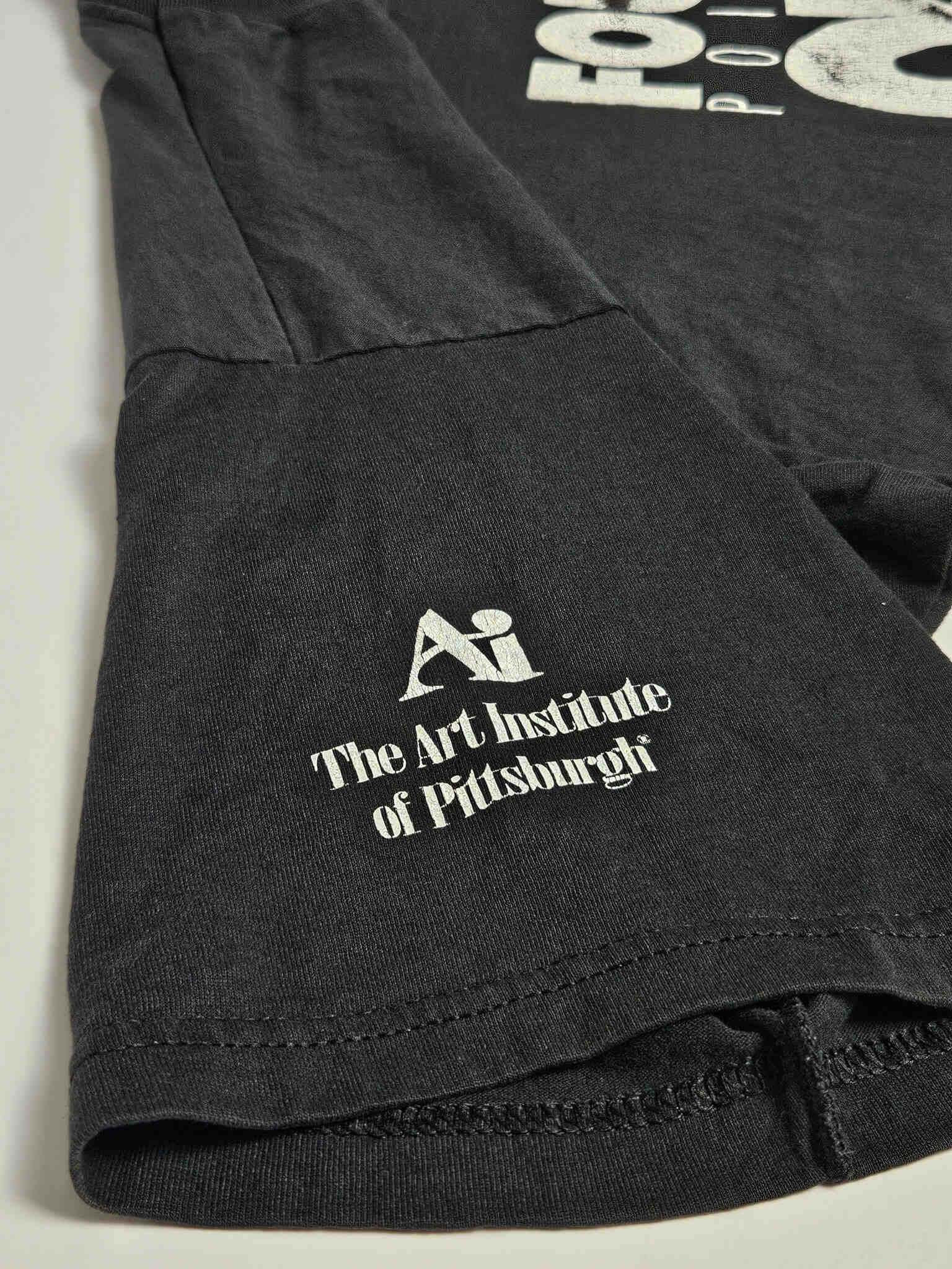 "Close-up van de mouw van een zwart vintage T-shirt, met een wit bedrukt logo van 'The Art Institute of Pittsburgh'. Dit detail voegt een unieke, historische context toe aan dit duurzame en authentieke slow fashion kledingstuk."

