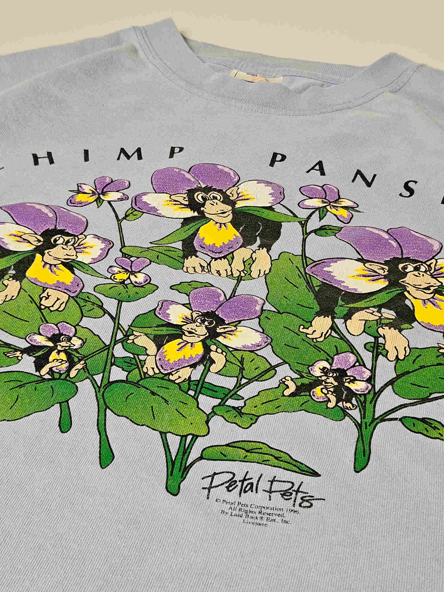 "Close-up van het bovenste gedeelte van een lichtblauw vintage T-shirt, waarop de 'CHIMP PANSIES' tekst en een deel van de grafische print te zien zijn. Onder de bloemen staat in kleine letters 'Petal Pets' gedrukt met een copyrightjaar 1999. Dit detail benadrukt de zeldzaamheid en het unieke karakter van dit vintage T-shirt, ideaal voor verzamelaars van duurzame mode."

