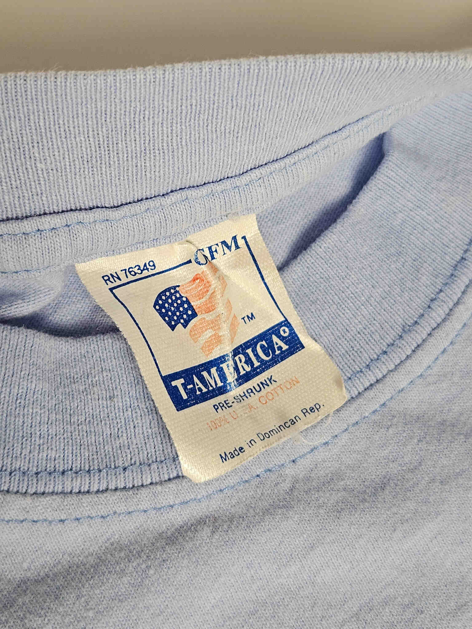 "Close-up van het witte textiellabel in de nek van een lichtblauw vintage T-shirt. Het label toont de tekst 'T-AMERICA' met een Amerikaanse vlag-afbeelding, en details zoals 'PRE-SHRUNK', '100% U.S.A. COTTON' en 'Made in Dominican Rep.'. Dit label bevestigt de authenticiteit en oorsprong van dit kwalitatieve slow fashion item."
