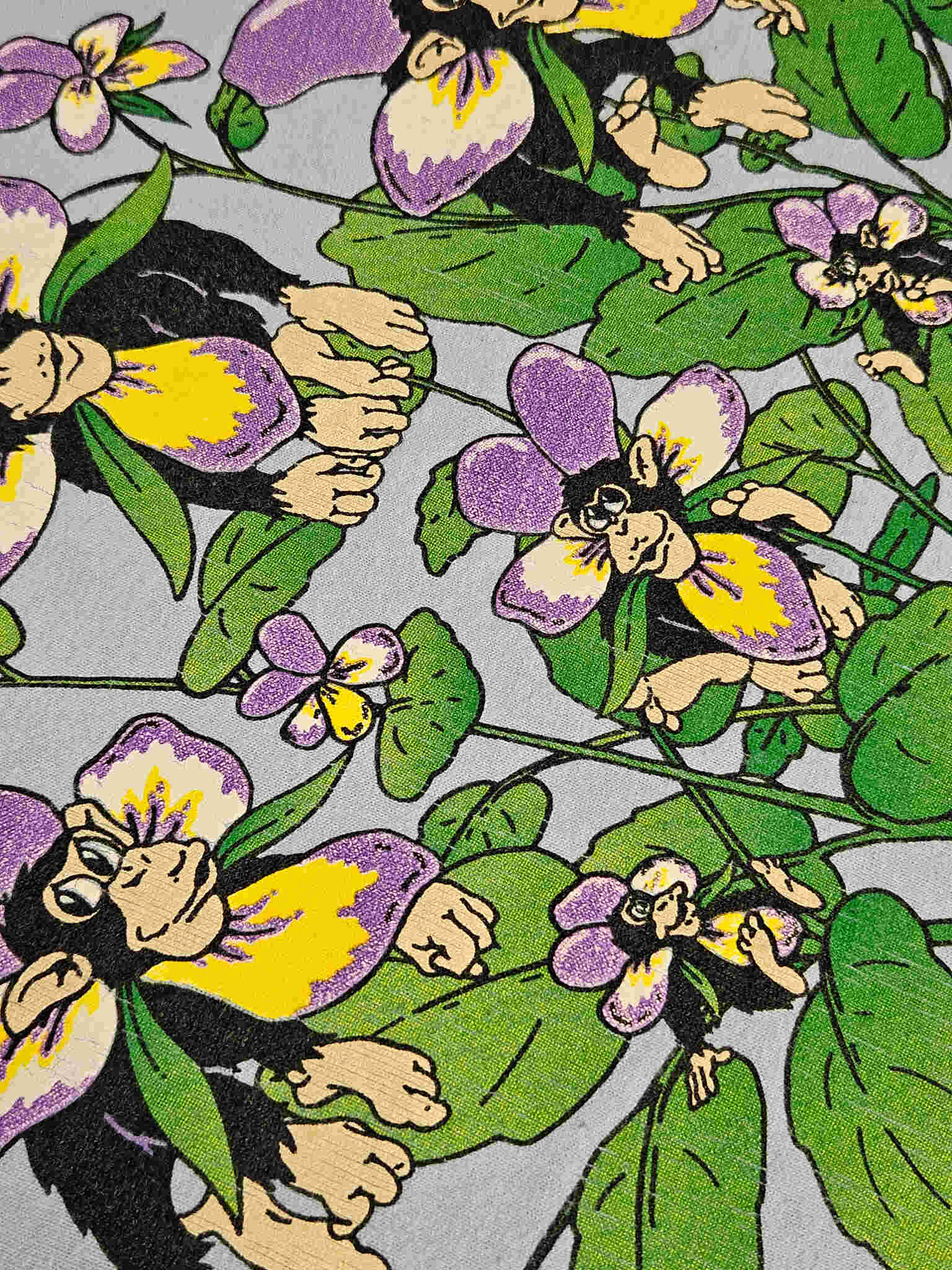 "Gedetailleerde close-up van de 'Chimp Pansies' print op een lichtblauw vintage T-shirt, waarbij de illustratie van chimpansees die uit paarse en gele viooltjes tevoorschijn komen, duidelijk zichtbaar is. De levendige kleuren en het unieke design benadrukken de charme van dit vintage kledingstuk."

