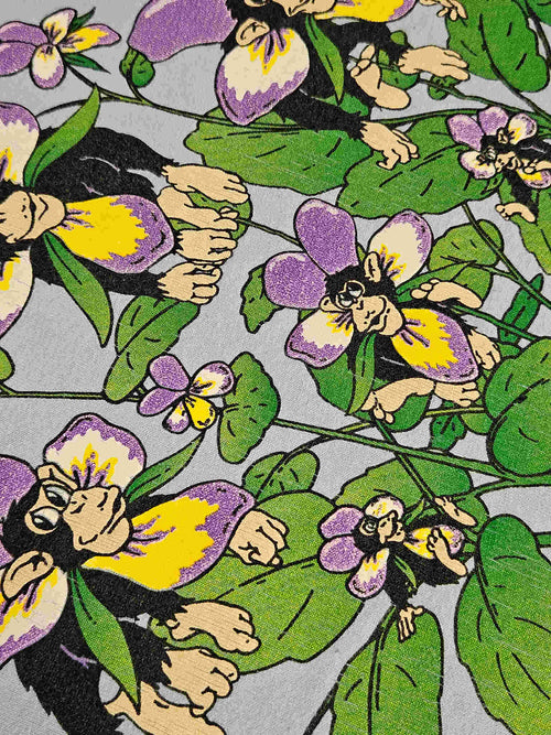 "Gedetailleerde close-up van de 'Chimp Pansies' print op een lichtblauw vintage T-shirt, waarbij de illustratie van chimpansees die uit paarse en gele viooltjes tevoorschijn komen, duidelijk zichtbaar is. De levendige kleuren en het unieke design benadrukken de charme van dit vintage kledingstuk."

