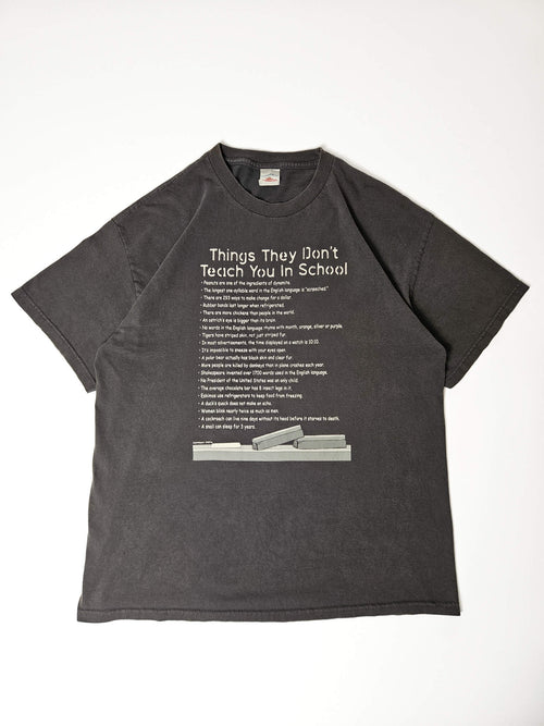 "Donkergrijs vintage T-shirt plat gelegd, met een humoristische witte print 'Things They Don't Teach You In School' bovenaan, gevolgd door een opsomming van verrassende feiten en onderaan een grafische weergave van gestapelde staven. Dit unieke en intrigerende item is een voorbeeld van duurzame, pre-loved slow fashion."


