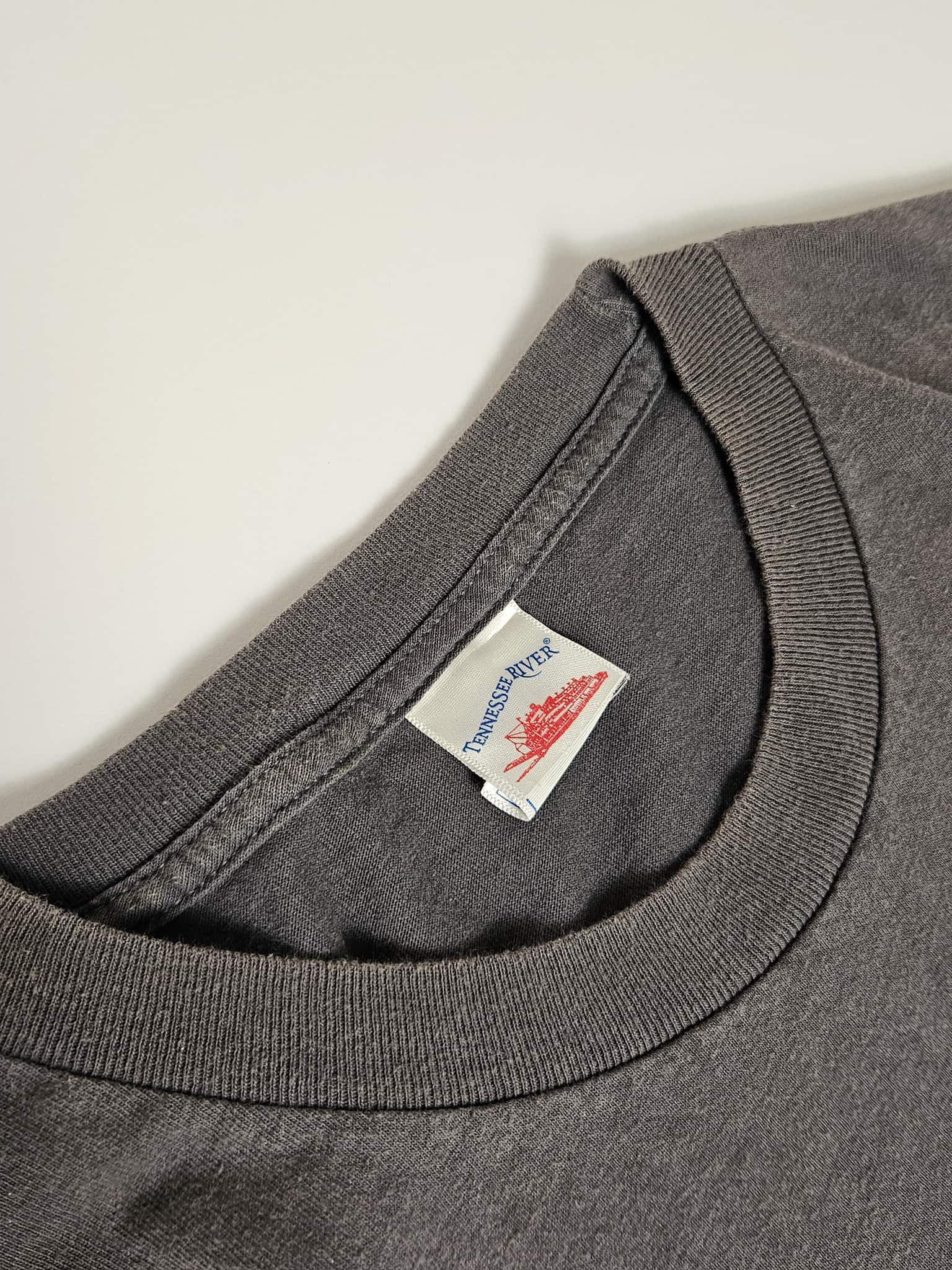 "Close-up van het witte label in de nek van een donkergrijs vintage T-shirt, waarop 'TENNESSEE RIVER' met een afbeelding van een rivierlandschap te zien is. Dit label helpt bij het authenticeren van de oorsprong en leeftijd van dit unieke slow fashion T-shirt."

