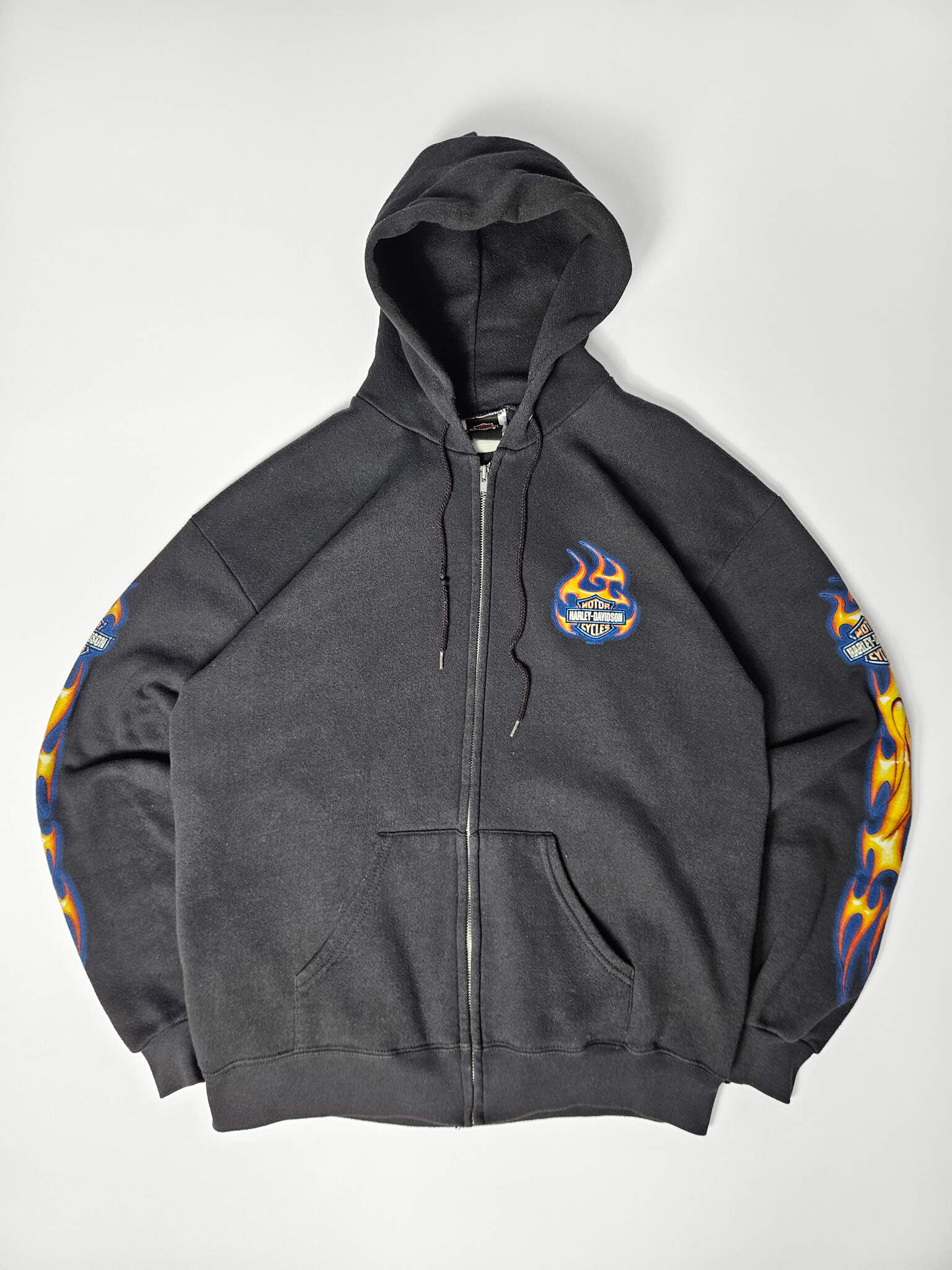 "Zwarte vintage Harley-Davidson zip-up hoodie, plat gelegd, met levendige blauwe en gele vlammenprints langs beide mouwen en een kleiner, vlammend Harley-Davidson logo op de linkerborst. Dit iconische en duurzame stuk is een must-have voor liefhebbers van authentieke motor- en slow fashion kleding."

