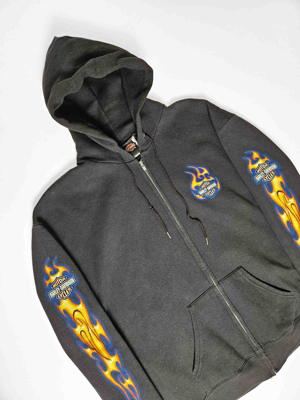 "Zwarte vintage Harley-Davidson zip-up hoodie, plat gelegd, met levendige blauwe en gele vlammenprints langs beide mouwen en een kleiner, vlammend Harley-Davidson logo op de linkerborst. Dit iconische en duurzame stuk is een must-have voor liefhebbers van authentieke motor- en slow fashion kleding."

