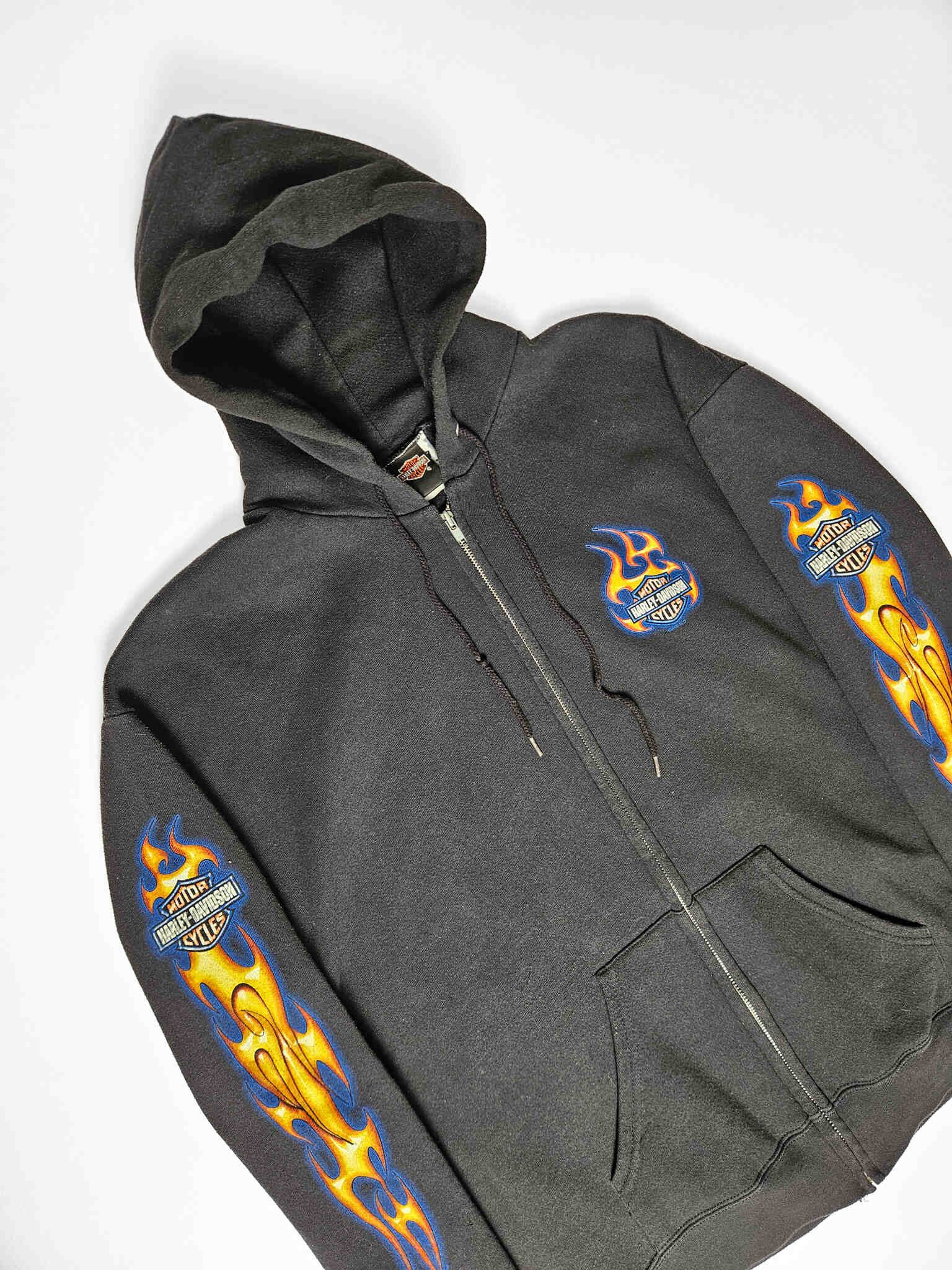 "Zwarte vintage Harley-Davidson zip-up hoodie, plat gelegd, met levendige blauwe en gele vlammenprints langs beide mouwen en een kleiner, vlammend Harley-Davidson logo op de linkerborst. Dit iconische en duurzame stuk is een must-have voor liefhebbers van authentieke motor- en slow fashion kleding."

