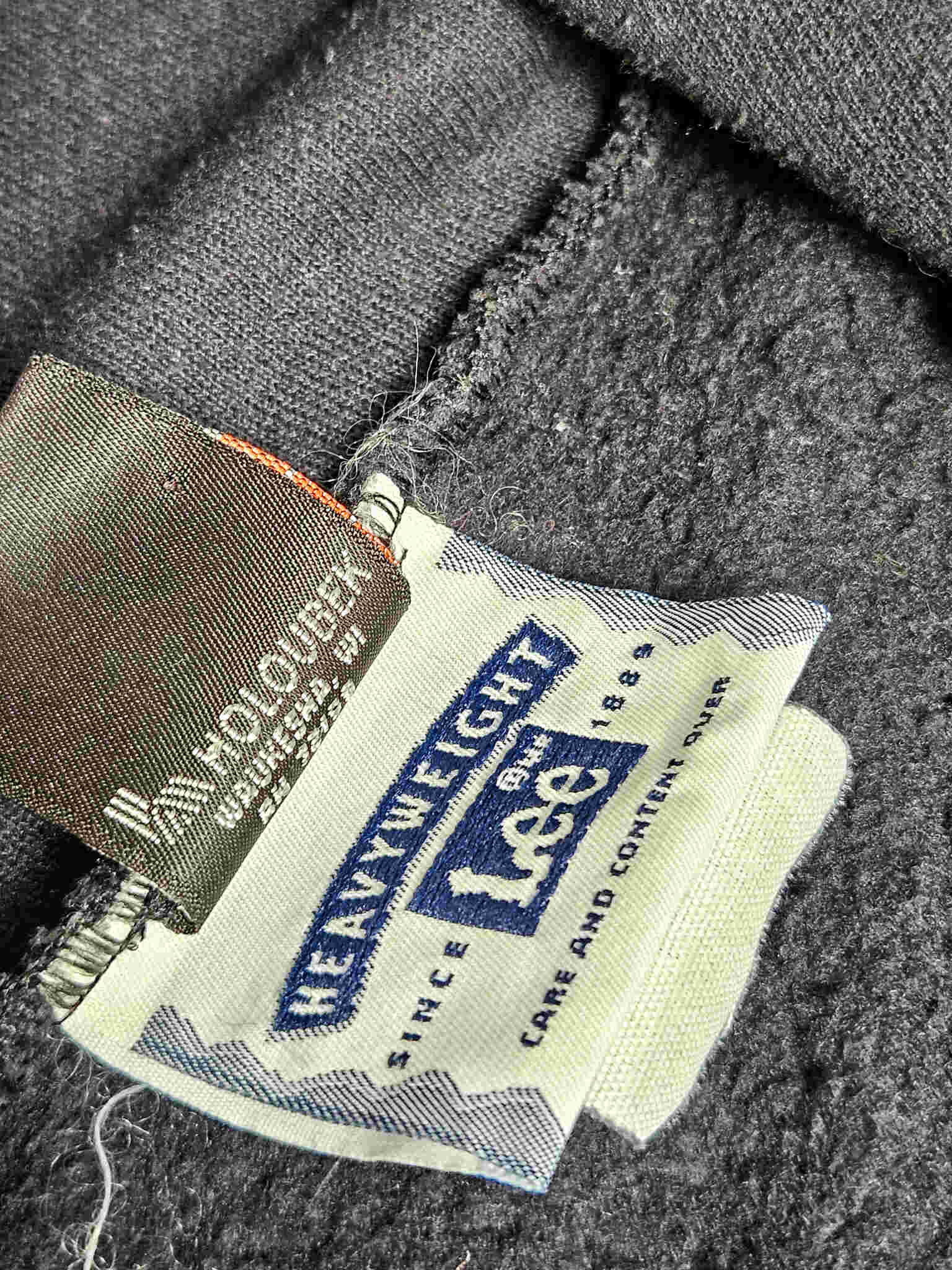 "Close-up van de interne labels van een zwarte vintage hoodie. Een wit label is duidelijk zichtbaar met 'HEAVYWEIGHT Lee' en 'SINCE 1889', wat duidt op de kwaliteit en leeftijd. Een ander zwart label met onleesbare tekst is erachter te zien. Dit detail bevestigt de authenticiteit van dit pre-loved slow fashion item."


