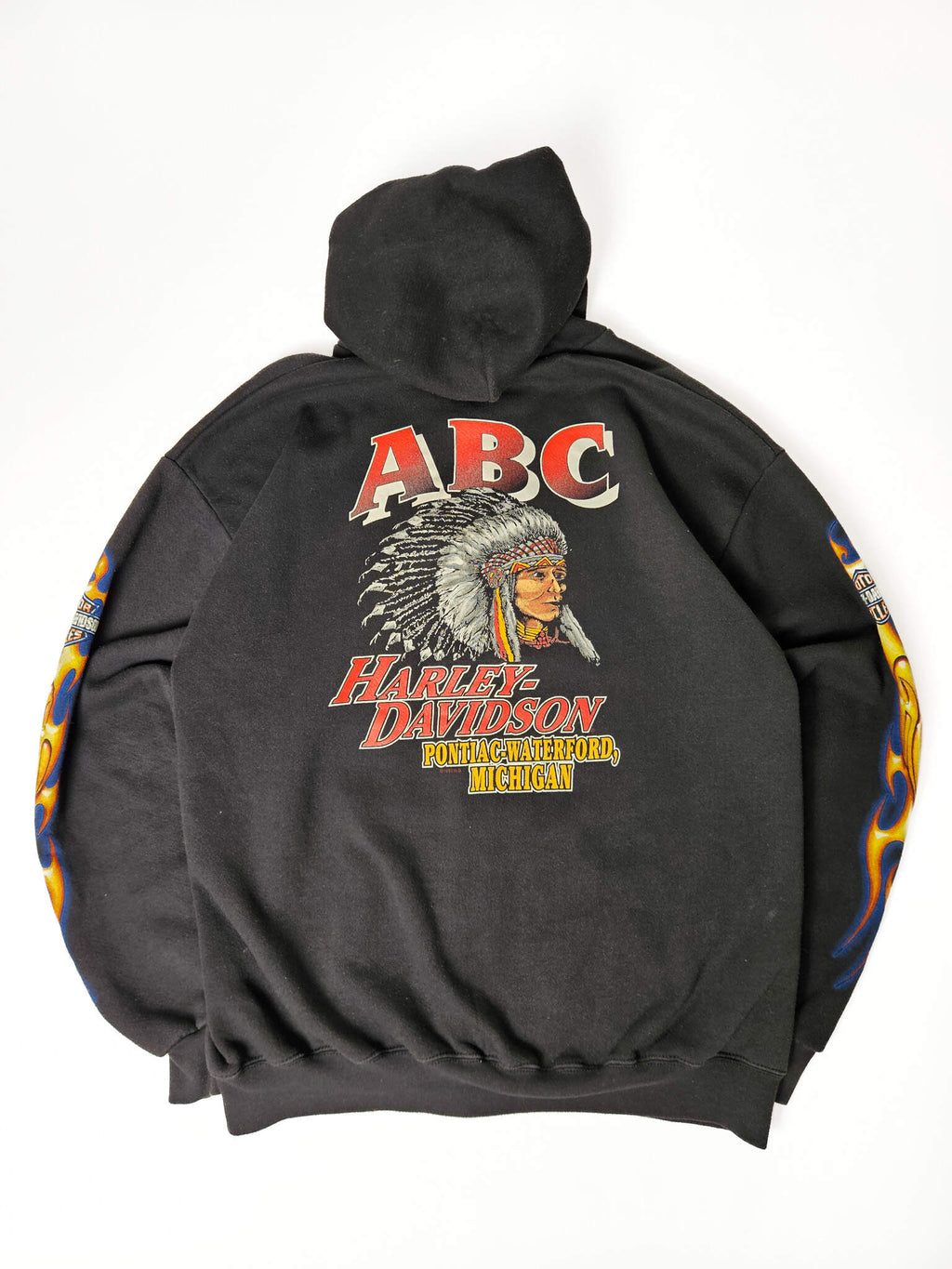 "Achterkant van een zwarte vintage Harley-Davidson zip-up hoodie, plat gelegd, met een grote opvallende print. Bovenaan staat 'ABC' in rood, daaronder een gedetailleerde illustratie van een Native American chief met hoofdtooien, gevolgd door 'HARLEY-DAVIDSON' en 'PONTIAC-WATERFORD, MICHIGAN'. Dit is een zeldzaam en krachtig stuk vintage motorwear, perfect voor slow fashion verzamelaars."

