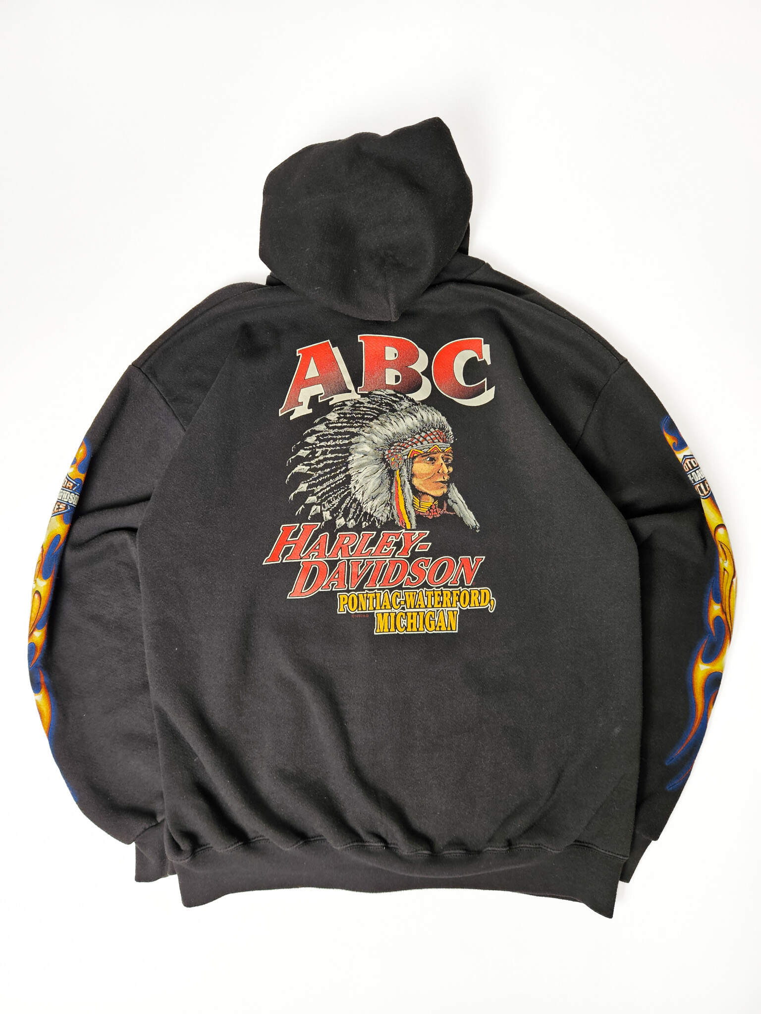"Achterkant van een zwarte vintage Harley-Davidson zip-up hoodie, plat gelegd, met een grote opvallende print. Bovenaan staat 'ABC' in rood, daaronder een gedetailleerde illustratie van een Native American chief met hoofdtooien, gevolgd door 'HARLEY-DAVIDSON' en 'PONTIAC-WATERFORD, MICHIGAN'. Dit is een zeldzaam en krachtig stuk vintage motorwear, perfect voor slow fashion verzamelaars."

