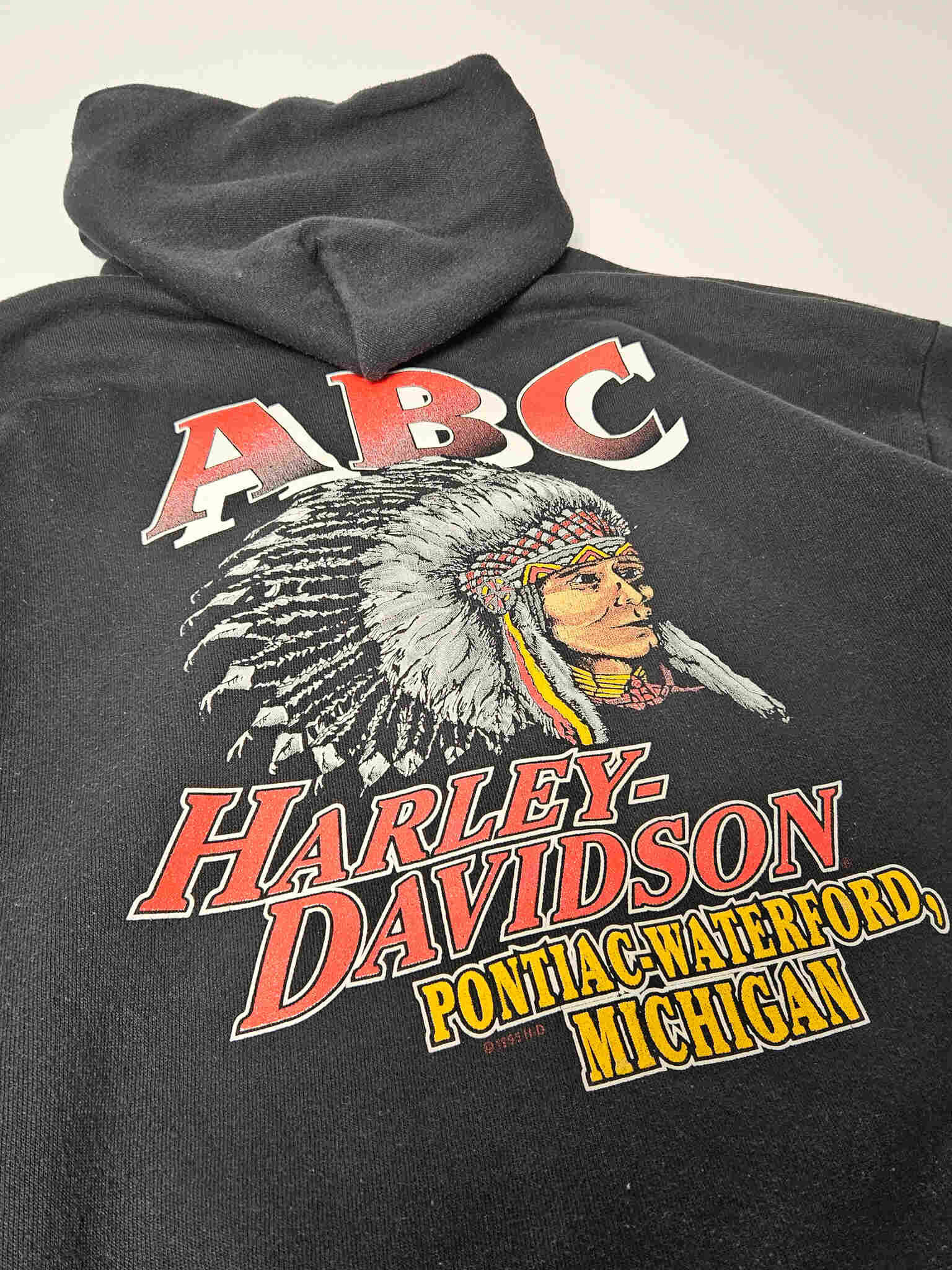 "Gedetailleerde close-up van de print op de achterkant van een zwarte vintage Harley-Davidson hoodie. De afbeelding toont het gezicht van een Native American chief met een rijk versierde hoofdtooien, erboven 'ABC' en eronder 'HARLEY-DAVIDSON PONTIAC-WATERFORD, MICHIGAN' in rode en gele letters. Deze vintage graphic is een sterk visueel element voor slow fashion liefhebbers."











