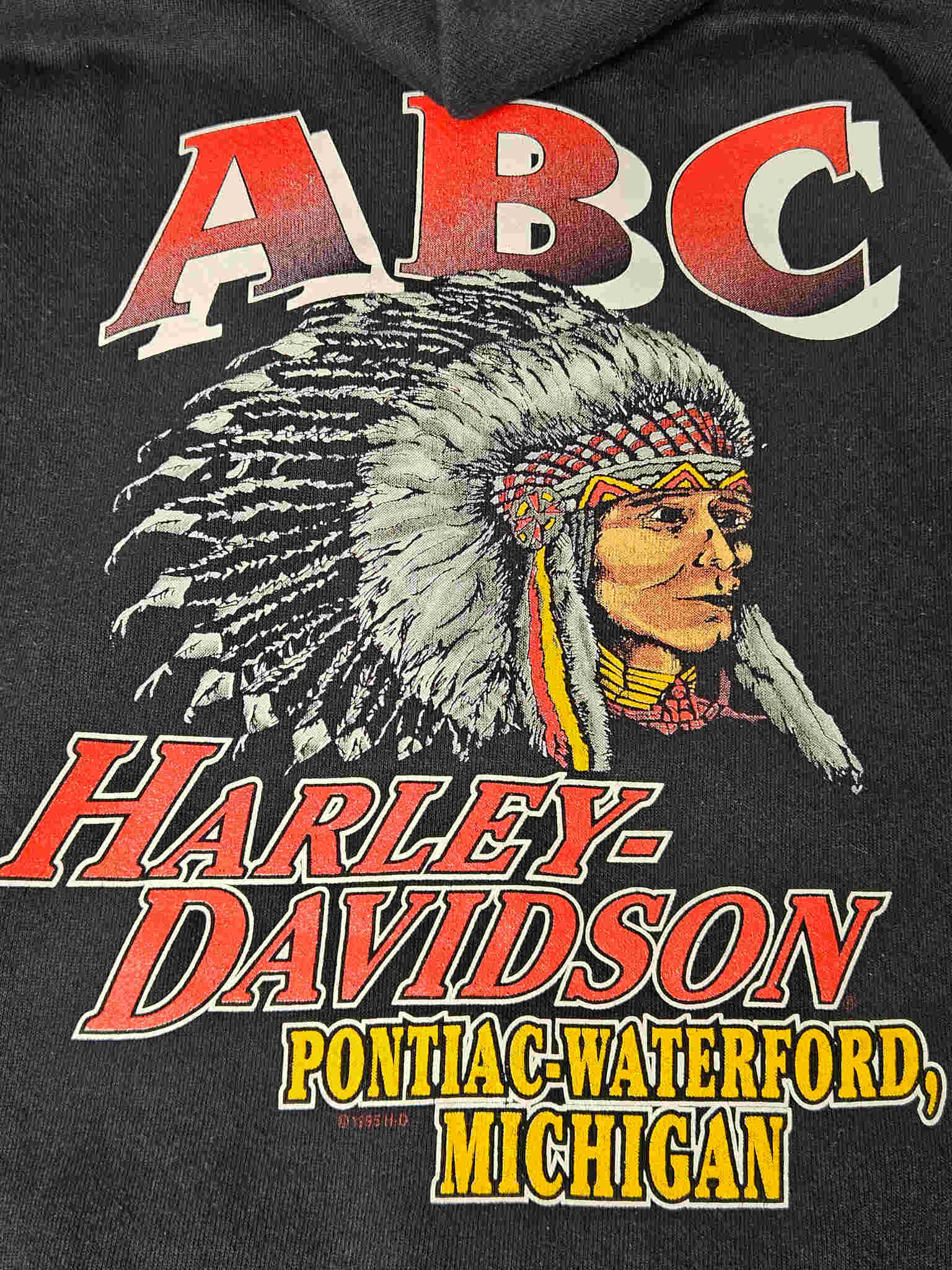 "Gedetailleerde close-up van de print op de achterkant van een zwarte vintage Harley-Davidson hoodie. De afbeelding toont het gezicht van een Native American chief met een rijk versierde hoofdtooien, erboven 'ABC' en eronder 'HARLEY-DAVIDSON PONTIAC-WATERFORD, MICHIGAN' in rode en gele letters. Deze vintage graphic is een sterk visueel element voor slow fashion liefhebbers."











