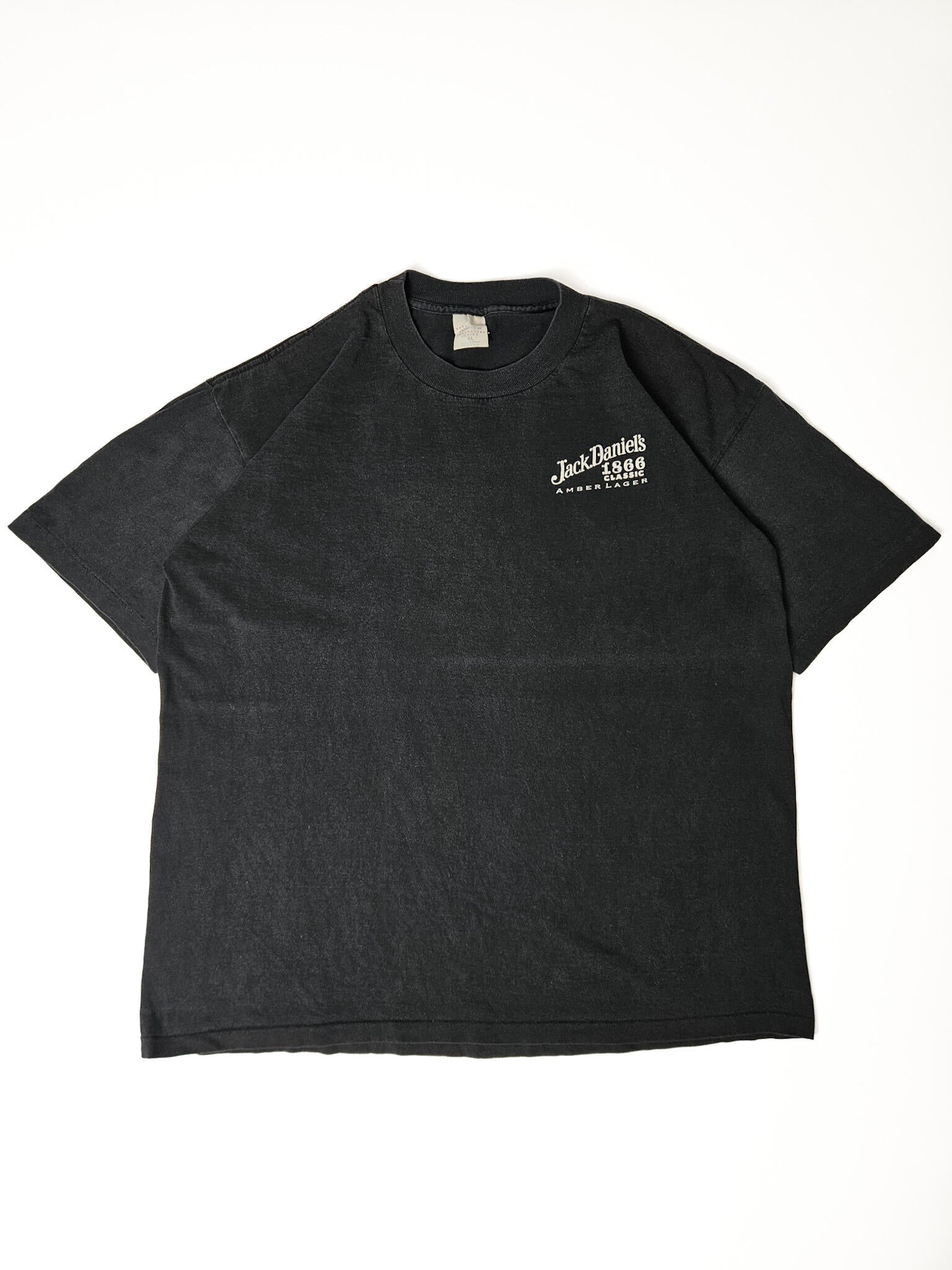 "Zwart, verweerd vintage T-shirt plat gelegd, met een klein wit logo van 'Jack Daniel's 1866 Classic Amber Lager' op de linkerborst. De subtiele print en het gedragen uiterlijk geven dit T-shirt een authentieke 'slow fashion' vintage uitstraling."

