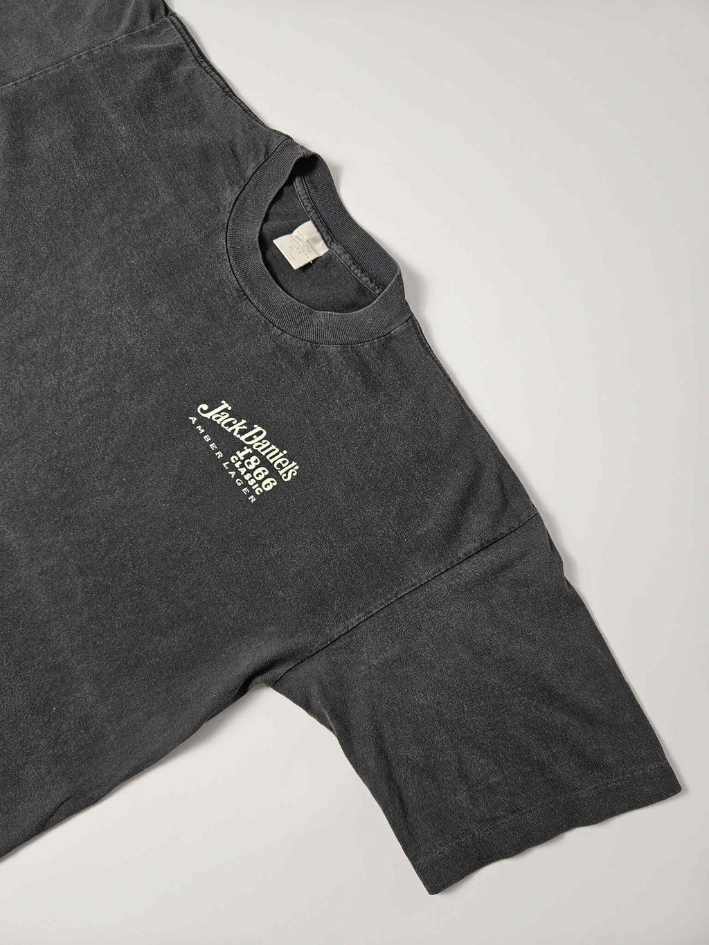 "Schuin perspectief van de voorkant van een zwart vintage T-shirt, waarop het kleine witte 'Jack Daniel's 1866 Classic Amber Lager' logo op de linkerborst zichtbaar is. De lichte vervaging van de stof benadrukt de vintage aard en de duurzaamheid van dit 'slow fashion' kledingstuk."

