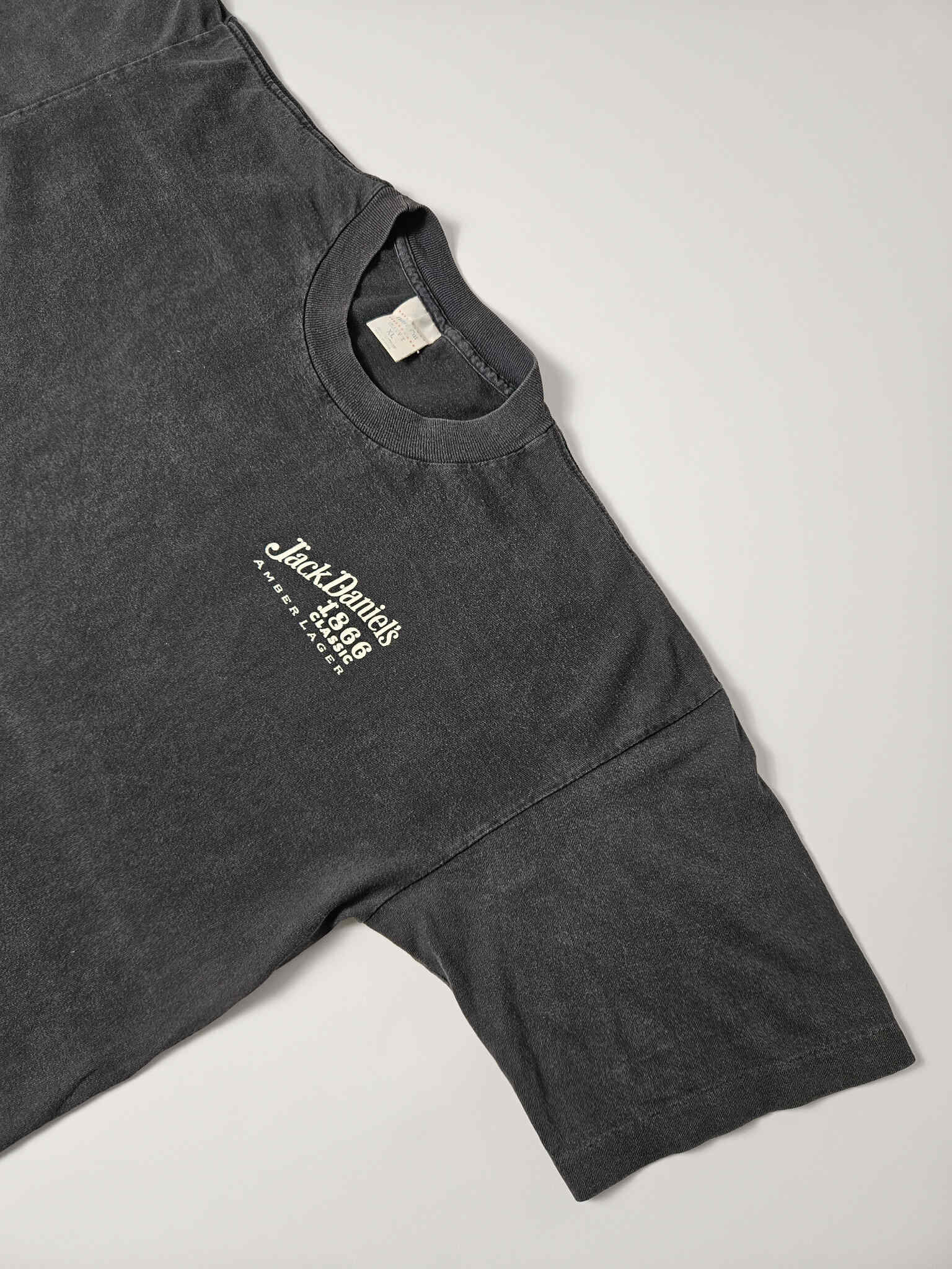 "Schuin perspectief van de voorkant van een zwart vintage T-shirt, waarop het kleine witte 'Jack Daniel's 1866 Classic Amber Lager' logo op de linkerborst zichtbaar is. De lichte vervaging van de stof benadrukt de vintage aard en de duurzaamheid van dit 'slow fashion' kledingstuk."

