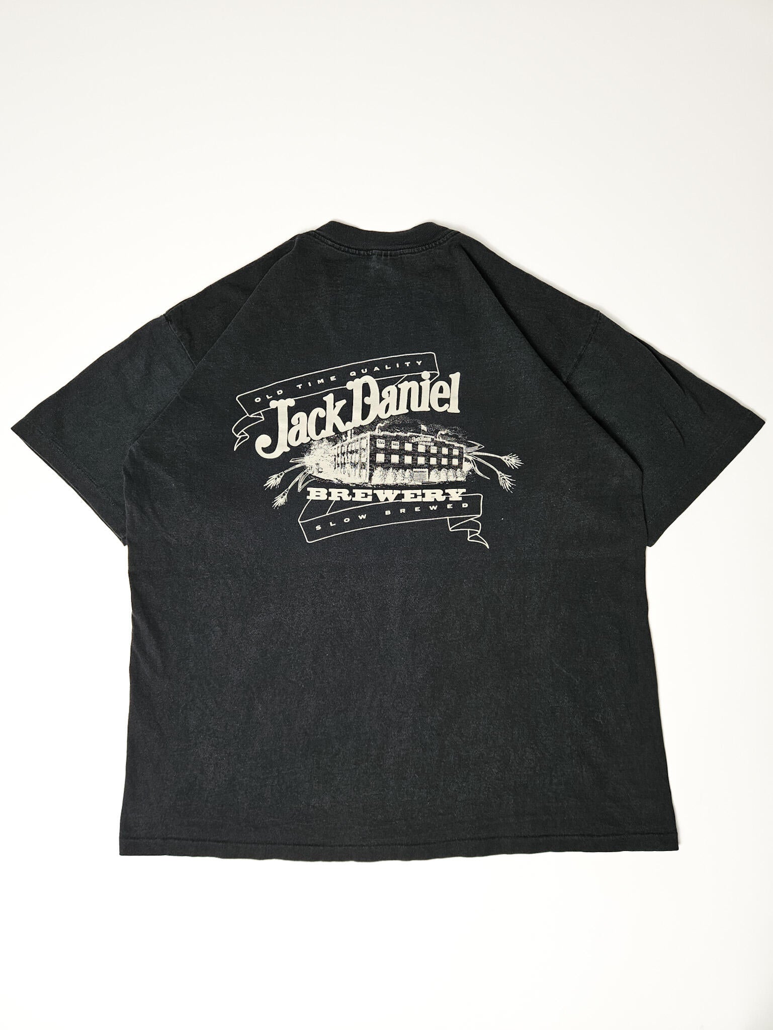 "Achterkant van een zwart vintage T-shirt plat gelegd, met een grote witte 'OLD TIME QUALITY Jack Daniel's BREWERY SLOW BREWED' print, inclusief een gedetailleerde illustratie van een gebouw. Dit indrukwekkende vintage T-shirt is een uitstekend voorbeeld van duurzame en unieke slow fashion kleding."










