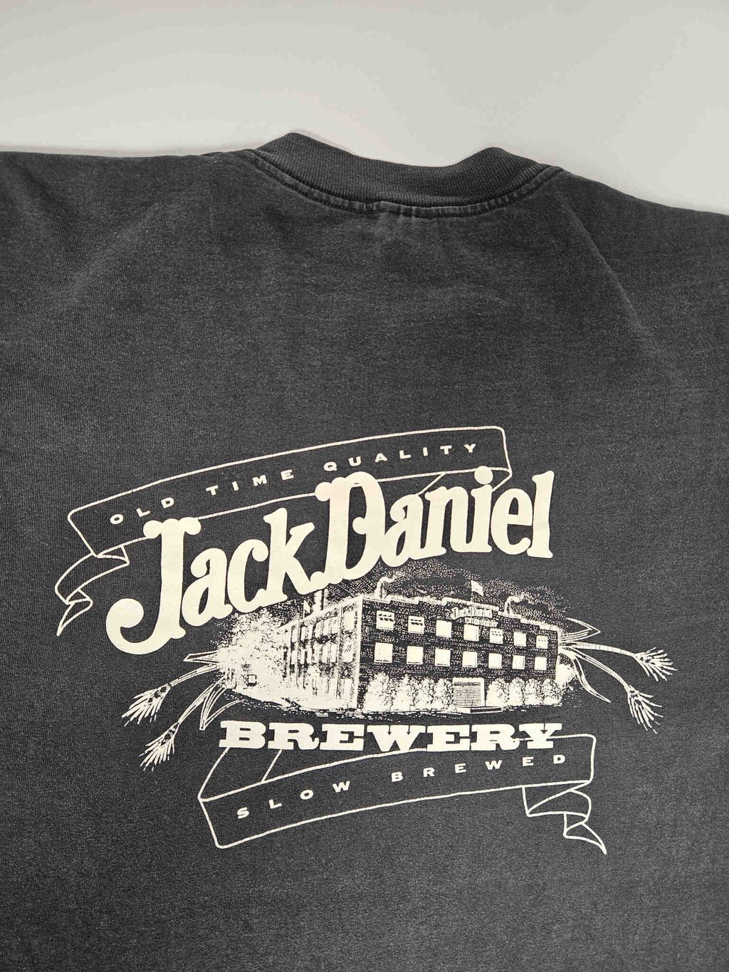 "Gedetailleerde close-up van de grote witte print op de achterkant van een zwart vintage T-shirt. De print toont 'OLD TIME QUALITY Jack Daniel's BREWERY SLOW BREWED' met een illustratie van een brouwerijgebouw en graanaren. Deze unieke grafische weergave draagt bij aan de zeldzaamheid van dit slow fashion verzamelstuk."

