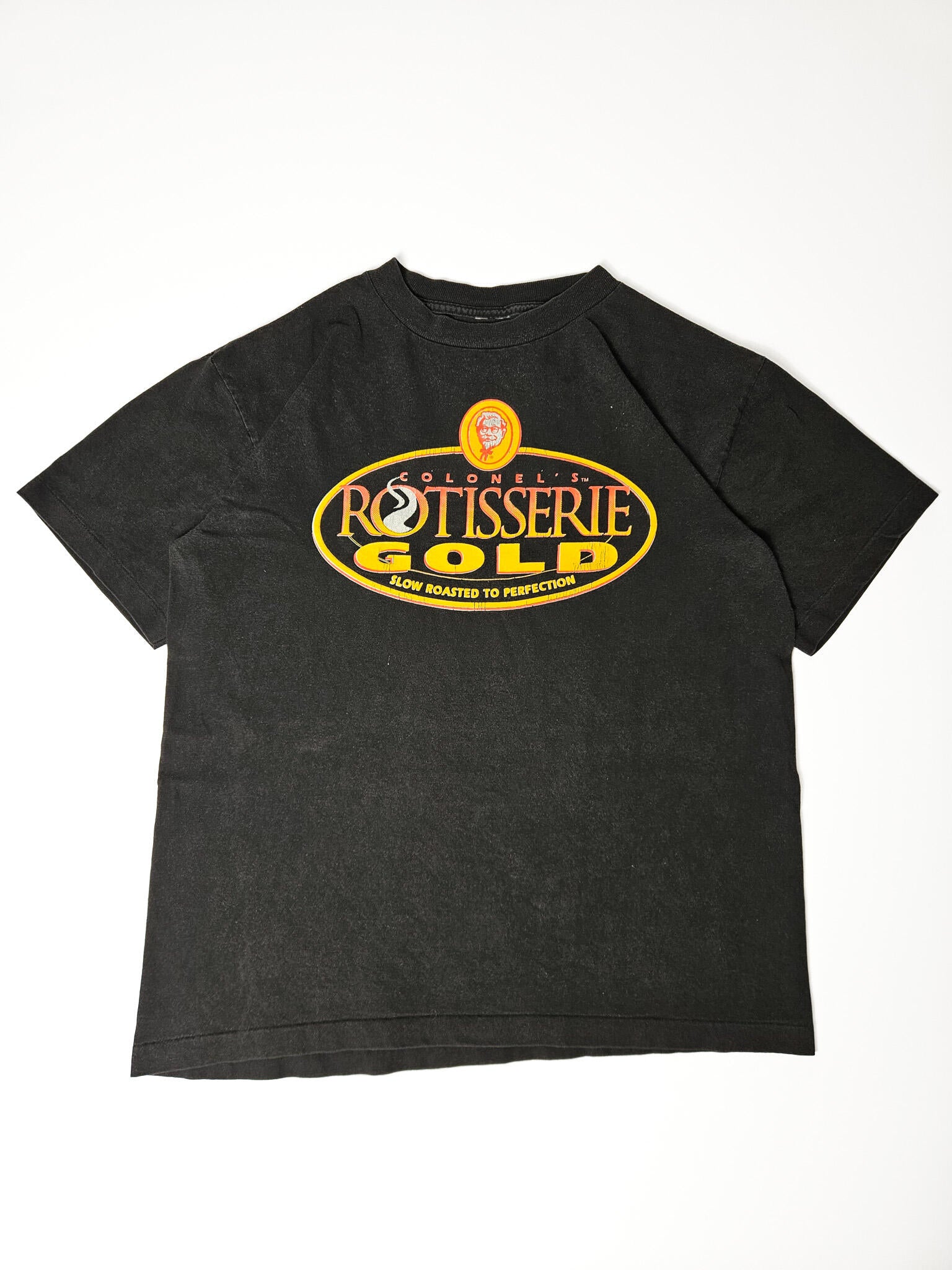"Zwart, verweerd vintage T-shirt plat gelegd, met een groot ovaal logo in rood en geel waarop 'COLONEL'S ROTISSERIE GOLD SLOW ROASTED TO PERFECTION' staat, inclusief een portret van Colonel Sanders. Dit iconische stuk is een unieke vondst en een perfect voorbeeld van duurzame 'slow fashion' kleding."

