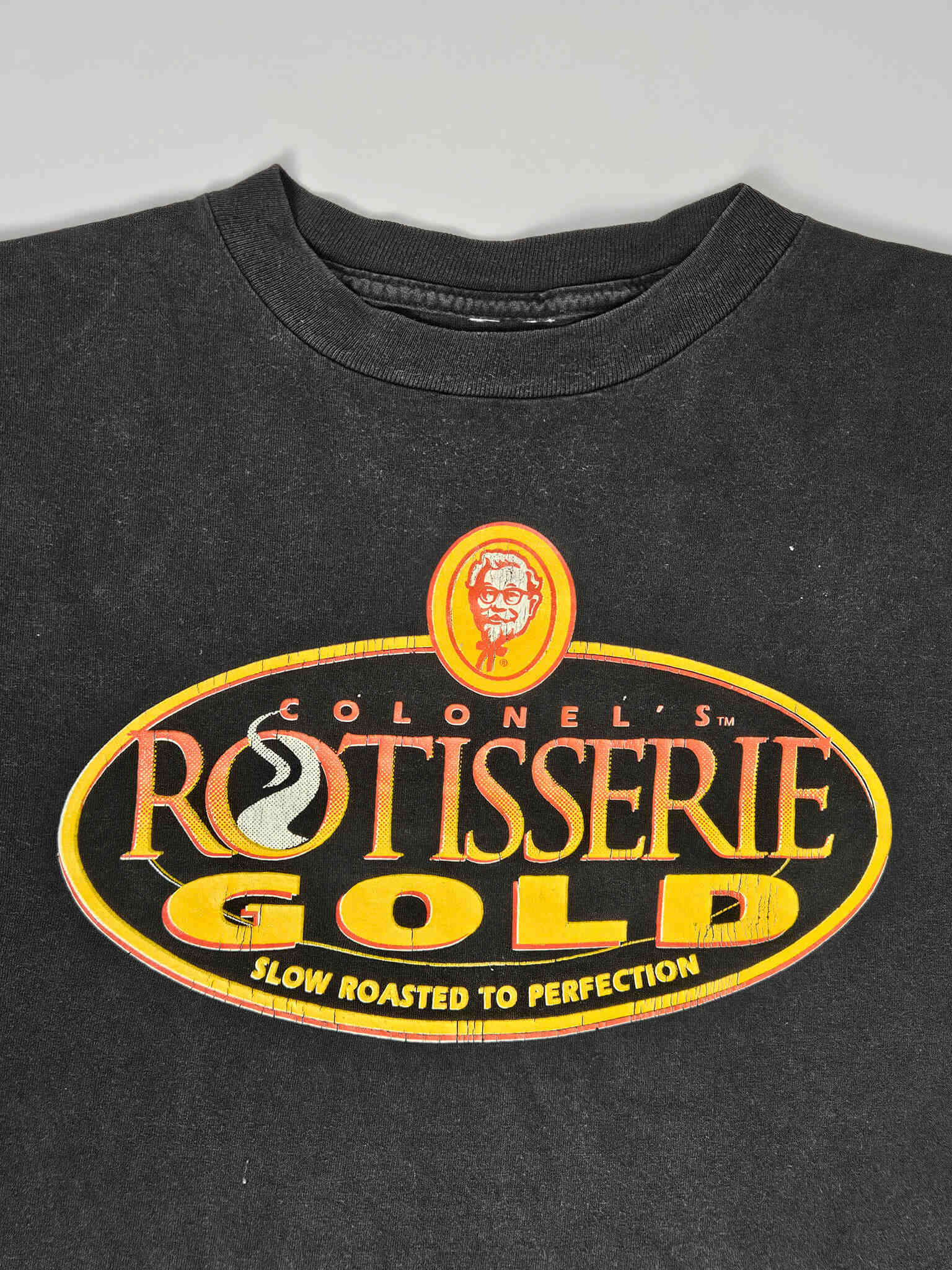 "Close-up van de bovenkant van een zwart vintage T-shirt, waarop het 'COLONEL'S ROTISSERIE GOLD' logo met het portret van Colonel Sanders prominent in beeld is. De lichte vervaging van de stof rond de nek en het logo benadrukt de authentieke vintage uitstraling van dit 'slow fashion' item."

