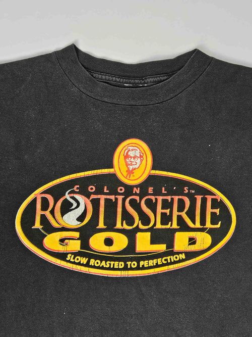 "Close-up van de bovenkant van een zwart vintage T-shirt, waarop het 'COLONEL'S ROTISSERIE GOLD' logo met het portret van Colonel Sanders prominent in beeld is. De lichte vervaging van de stof rond de nek en het logo benadrukt de authentieke vintage uitstraling van dit 'slow fashion' item."

