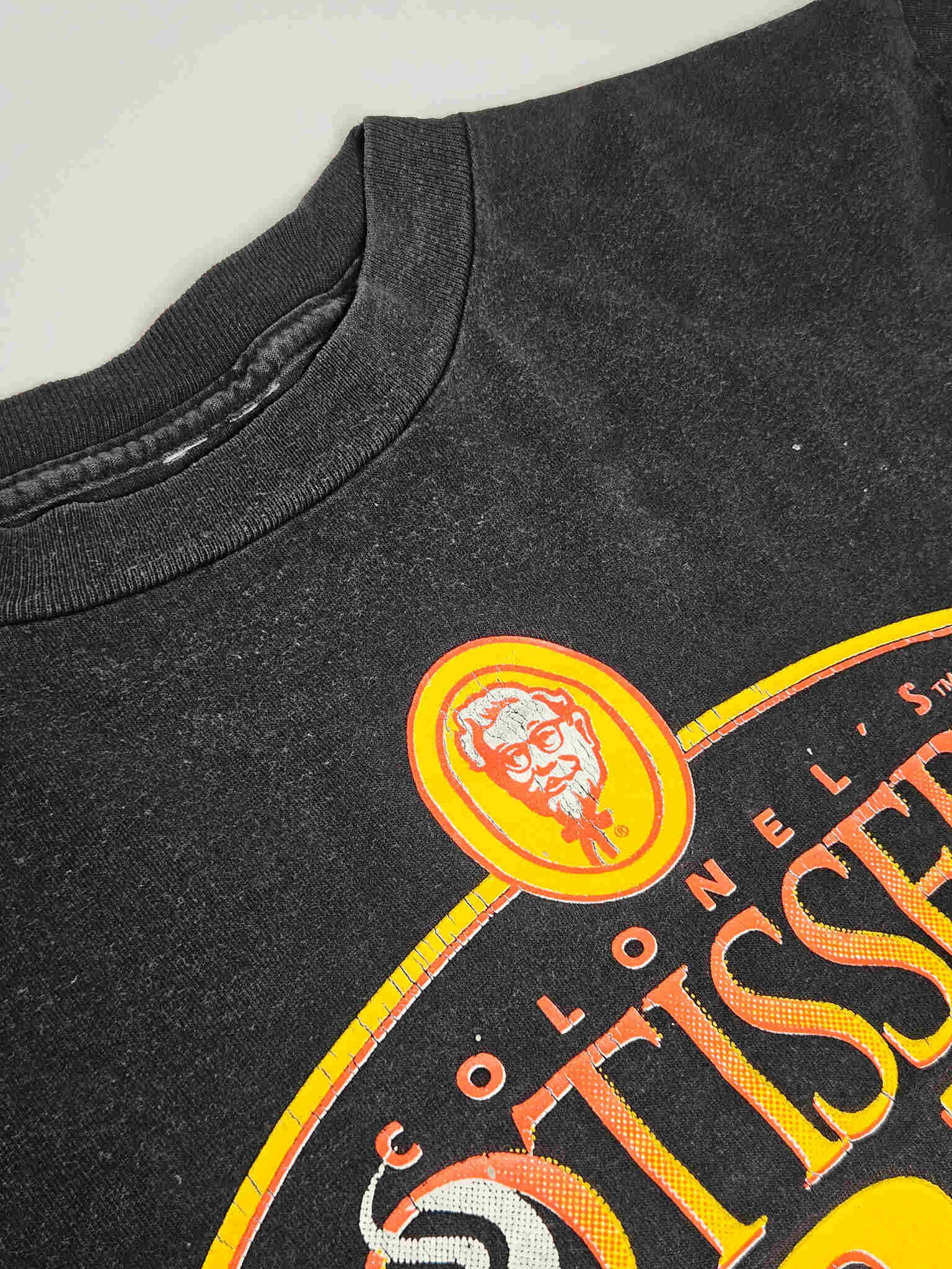"Close-up van de bovenkant van een zwart vintage T-shirt, waarop het 'COLONEL'S ROTISSERIE GOLD' logo met het portret van Colonel Sanders prominent in beeld is. De lichte vervaging van de stof rond de nek en het logo benadrukt de authentieke vintage uitstraling van dit 'slow fashion' item."

