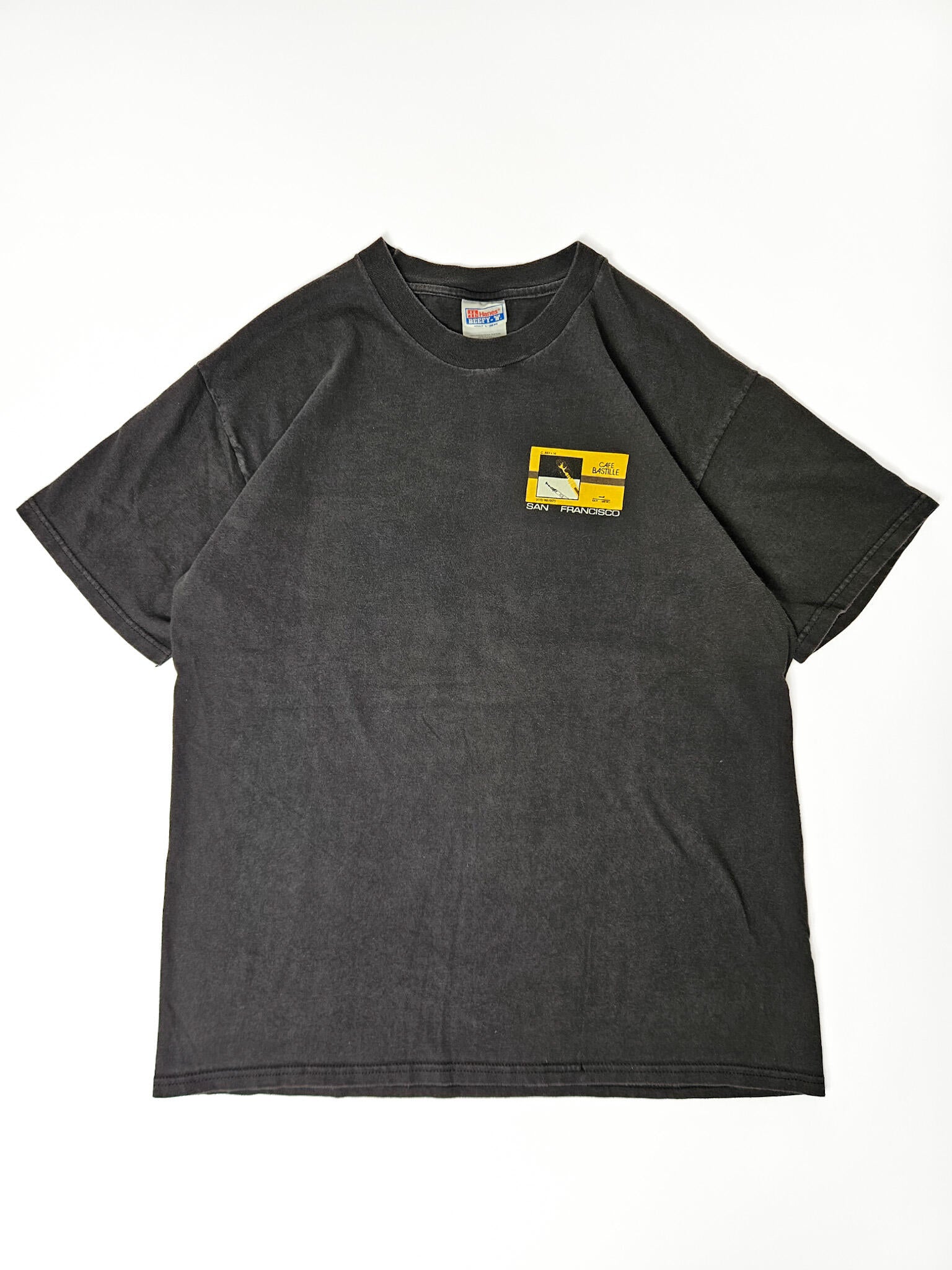 "Zwart, verweerd vintage T-shirt plat gelegd, met een klein vierkant logo op de linkerborst in geel en zwart, met een abstracte afbeelding en de tekst 'SAN FRANCISCO'. Dit subtiele en tijdloze 'slow fashion' T-shirt draagt de charme van vintage reissouvenirs."


