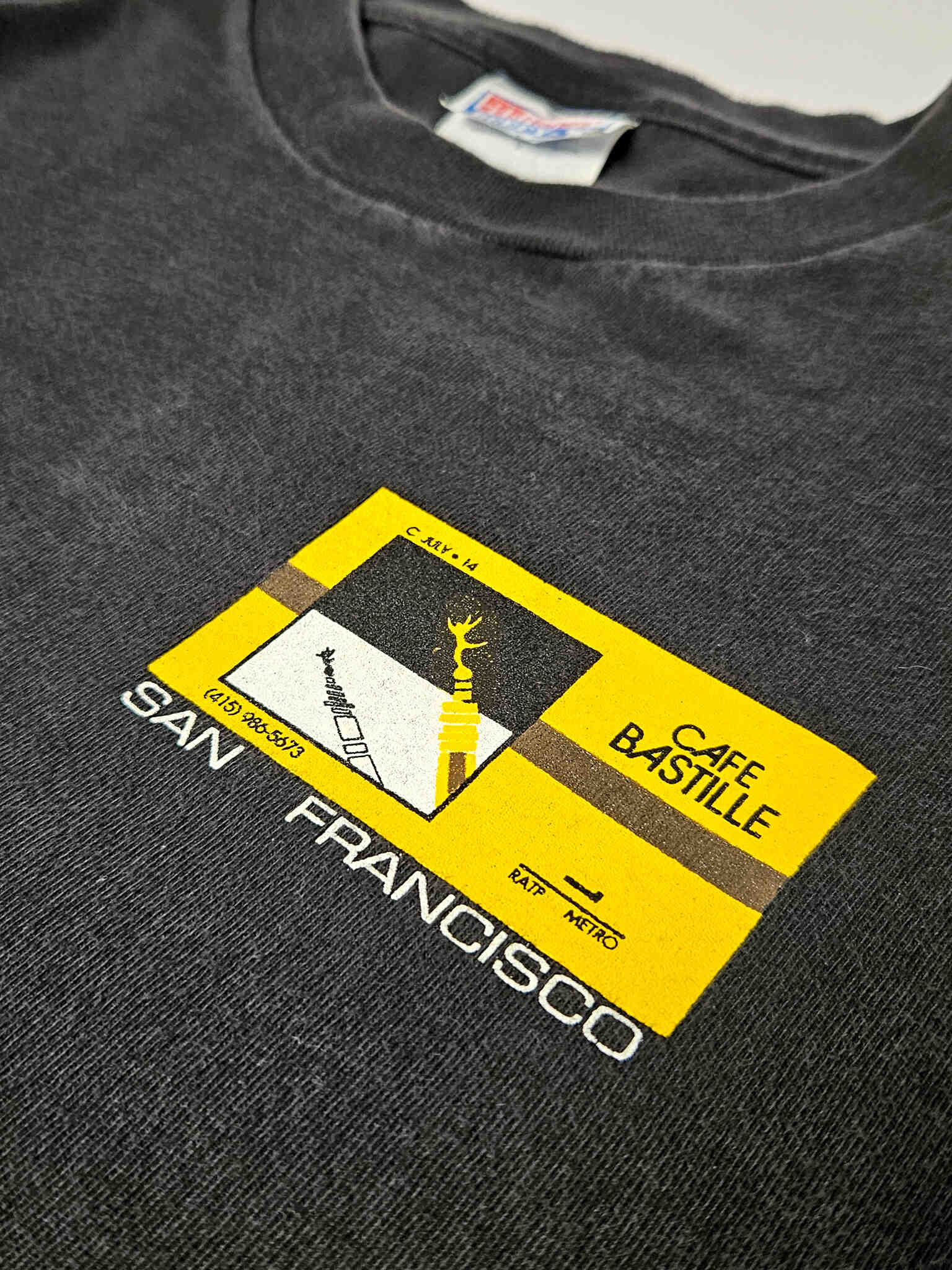 "Gedetailleerde close-up van het kleine vierkante logo op de linkerborst van een zwart vintage T-shirt. Het geel-zwarte design bevat abstracte vormen en de tekst 'SAN FRANCISCO', wat de herkomst van dit authentieke en duurzame 'slow fashion' kledingstuk aangeeft."

