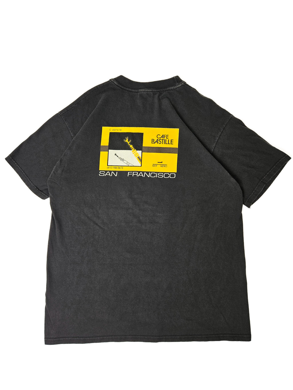 "Achterkant van een zwart vintage T-shirt plat gelegd, met een groot rechthoekig geel en zwart grafisch design. De print bevat architectonische elementen, de tekst 'CAFE BASTILLE', 'SAN FRANCISCO', en een telefoonnummer. Dit unieke 'slow fashion' item vertelt een verhaal en is ideaal voor liefhebbers van vintage design."

