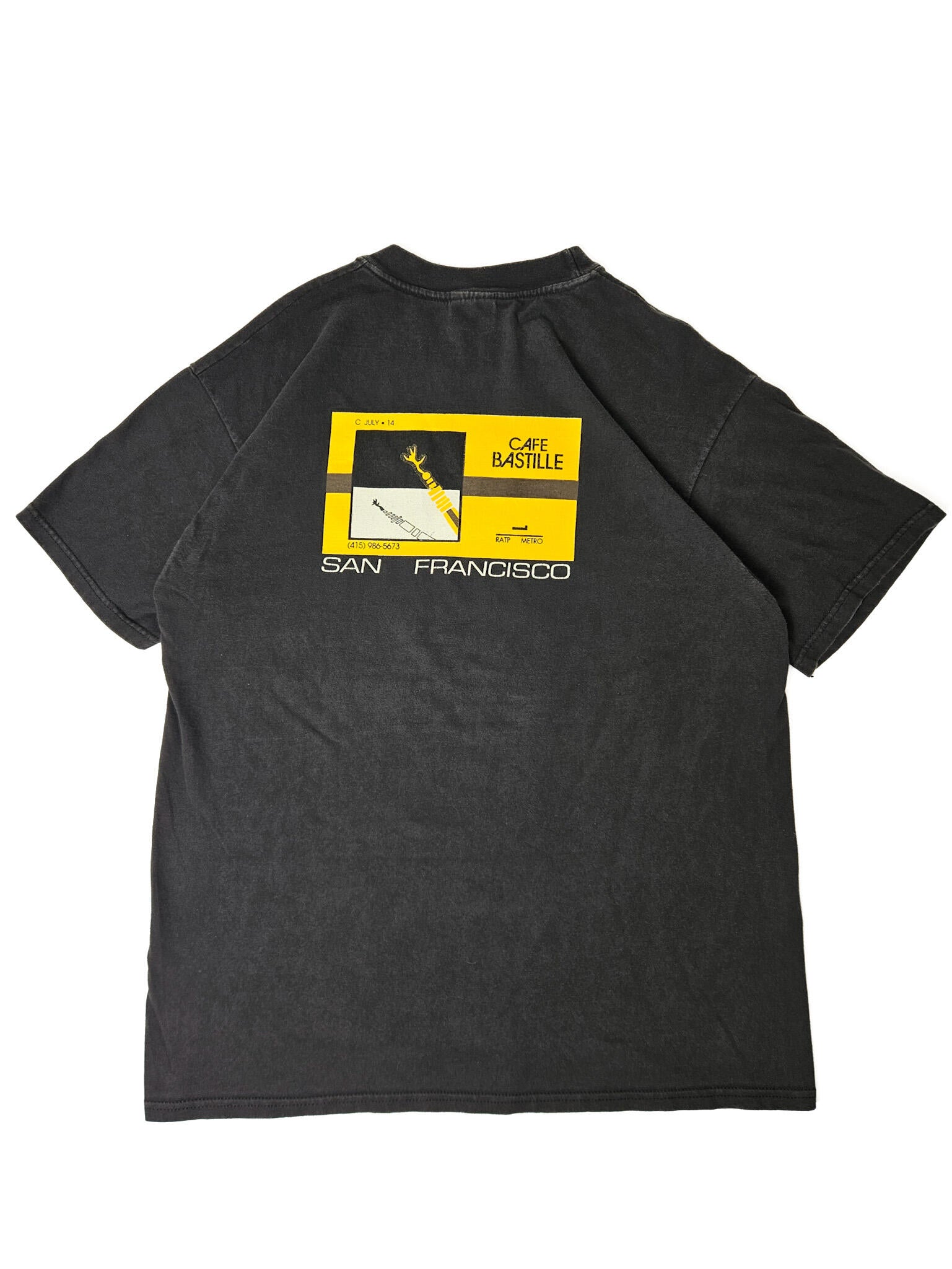 "Achterkant van een zwart vintage T-shirt plat gelegd, met een groot rechthoekig geel en zwart grafisch design. De print bevat architectonische elementen, de tekst 'CAFE BASTILLE', 'SAN FRANCISCO', en een telefoonnummer. Dit unieke 'slow fashion' item vertelt een verhaal en is ideaal voor liefhebbers van vintage design."

