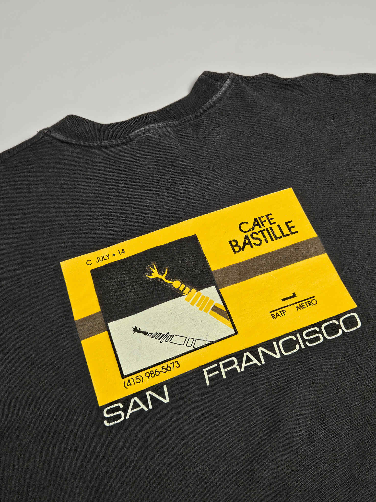 "Close-up van de bovenkant van het grote grafische design op de achterkant van een zwart vintage T-shirt. De gele en zwarte print toont 'CAFE BASTILLE' en abstracte architectonische vormen. De lichte vervaging van de print draagt bij aan de 'vintage' look van dit 'slow fashion' collector's item."

