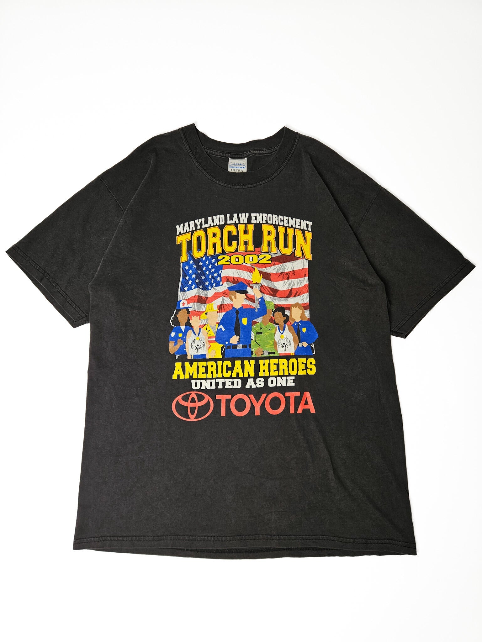 "Zwart, verweerd vintage T-shirt plat gelegd, met een grote meerkleurige print van 'MARYLAND LAW ENFORCEMENT TORCH RUN 2002'. De graphic toont diverse figuren (politie, brandweer, militairen) voor een Amerikaanse vlag, met de tekst 'AMERICAN HEROES UNITED AS ONE' en een rood Toyota-logo. Dit unieke en historische item is een perfect voorbeeld van duurzame 'slow fashion' evenementenkleding."