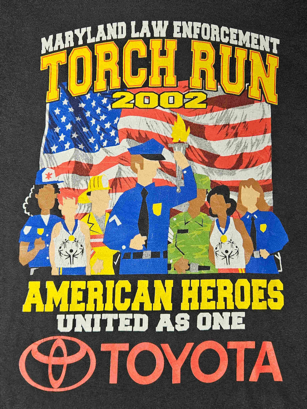 "Gedetailleerde close-up van de 'MARYLAND LAW ENFORCEMENT TORCH RUN 2002' print op een zwart vintage T-shirt. De afbeelding toont diverse beroepsgroepen voor een Amerikaanse vlag, met duidelijke tekst 'AMERICAN HEROES UNITED AS ONE' en het Toyota-logo. De details van de print en de datum 2002 maken dit een uniek verzamelstuk binnen de slow fashion categorie."