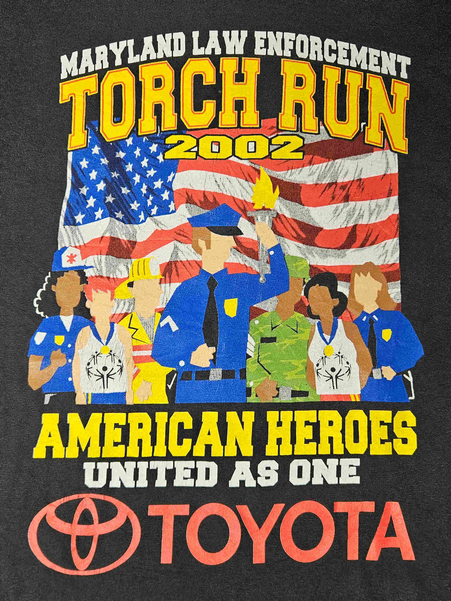 "Gedetailleerde close-up van de 'MARYLAND LAW ENFORCEMENT TORCH RUN 2002' print op een zwart vintage T-shirt. De afbeelding toont diverse beroepsgroepen voor een Amerikaanse vlag, met duidelijke tekst 'AMERICAN HEROES UNITED AS ONE' en het Toyota-logo. De details van de print en de datum 2002 maken dit een uniek verzamelstuk binnen de slow fashion categorie."
