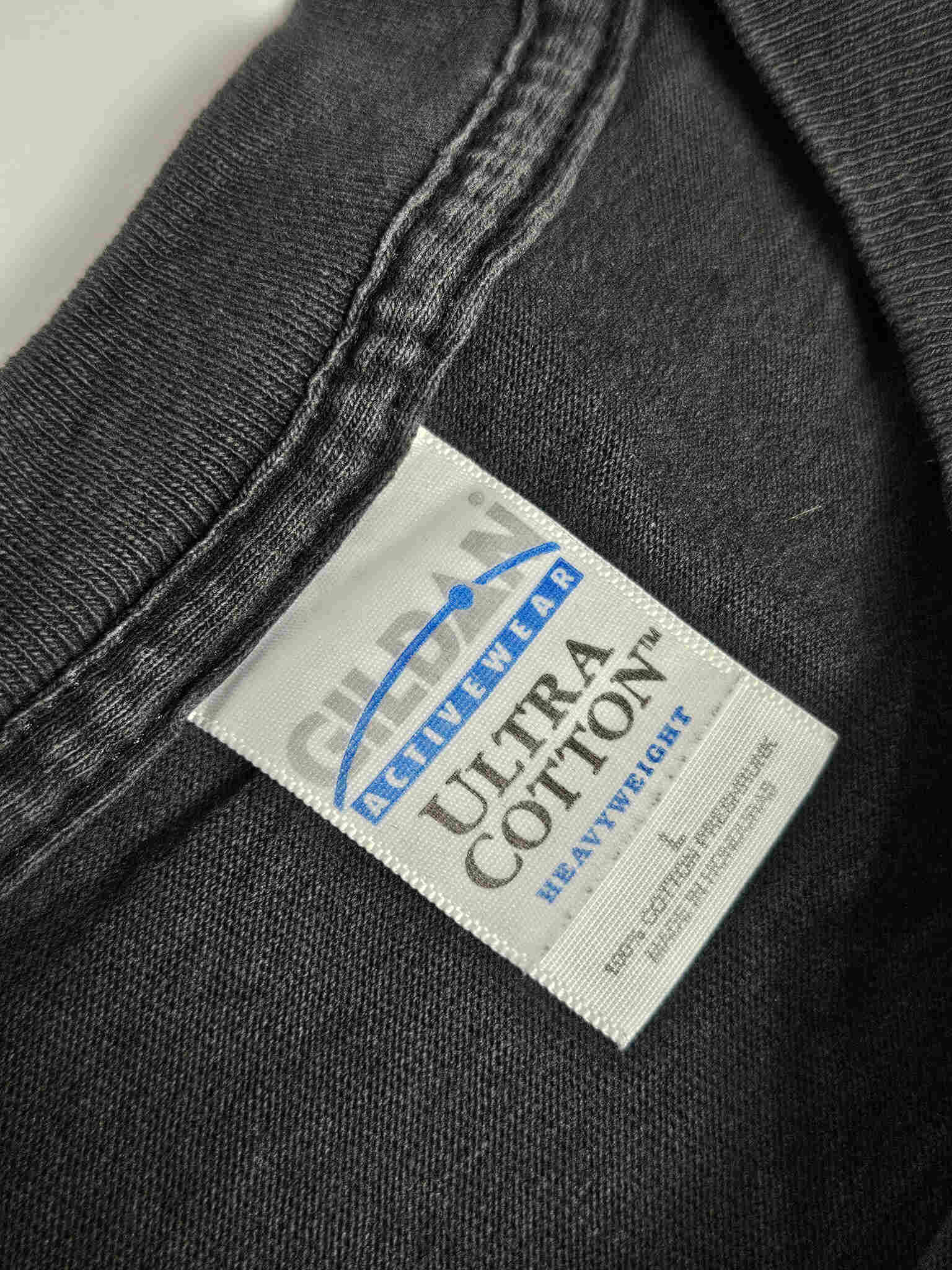 "Close-up van het witte label in de nek van een zwart vintage T-shirt, waarop 'GILDAN ACTIVEWEAR ULTRA COTTON HEAVYWEIGHT L' staat, samen met de tekst '100% COTTON PRE-SHRUNK MADE IN HONDURAS'. Dit label bevestigt de authenticiteit en de goede kwaliteit van dit 'slow fashion' T-shirt."