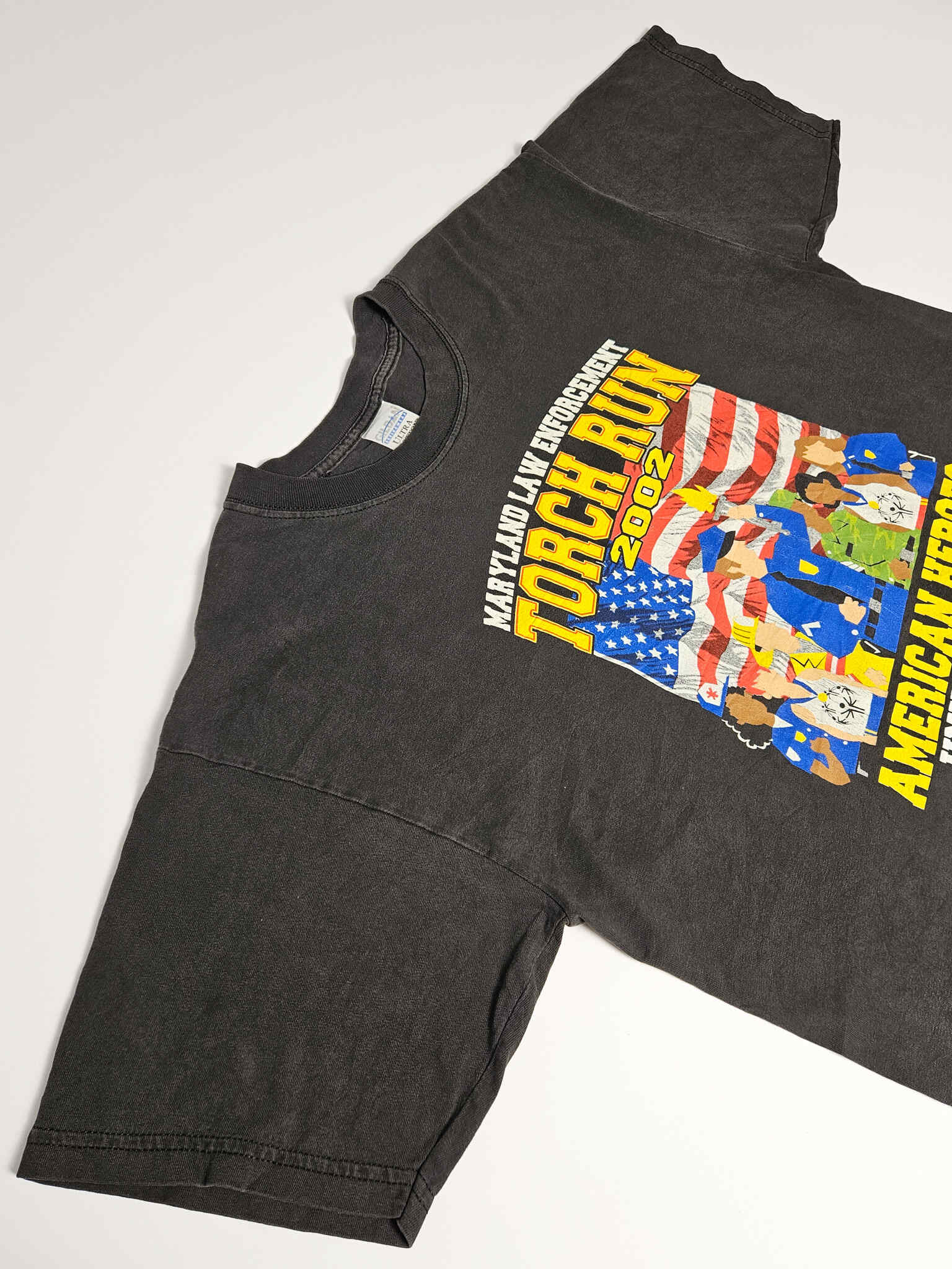 "Schuin perspectief van een zwart vintage T-shirt, waarop de grote 'MARYLAND LAW ENFORCEMENT TORCH RUN 2002' print duidelijk zichtbaar is. De lichte vervaging van de stof en de levendigheid van de kleuren zijn goed te zien. Dit authentieke evenementen T-shirt is een duurzame aanwinst voor de 'slow fashion' garderobe."